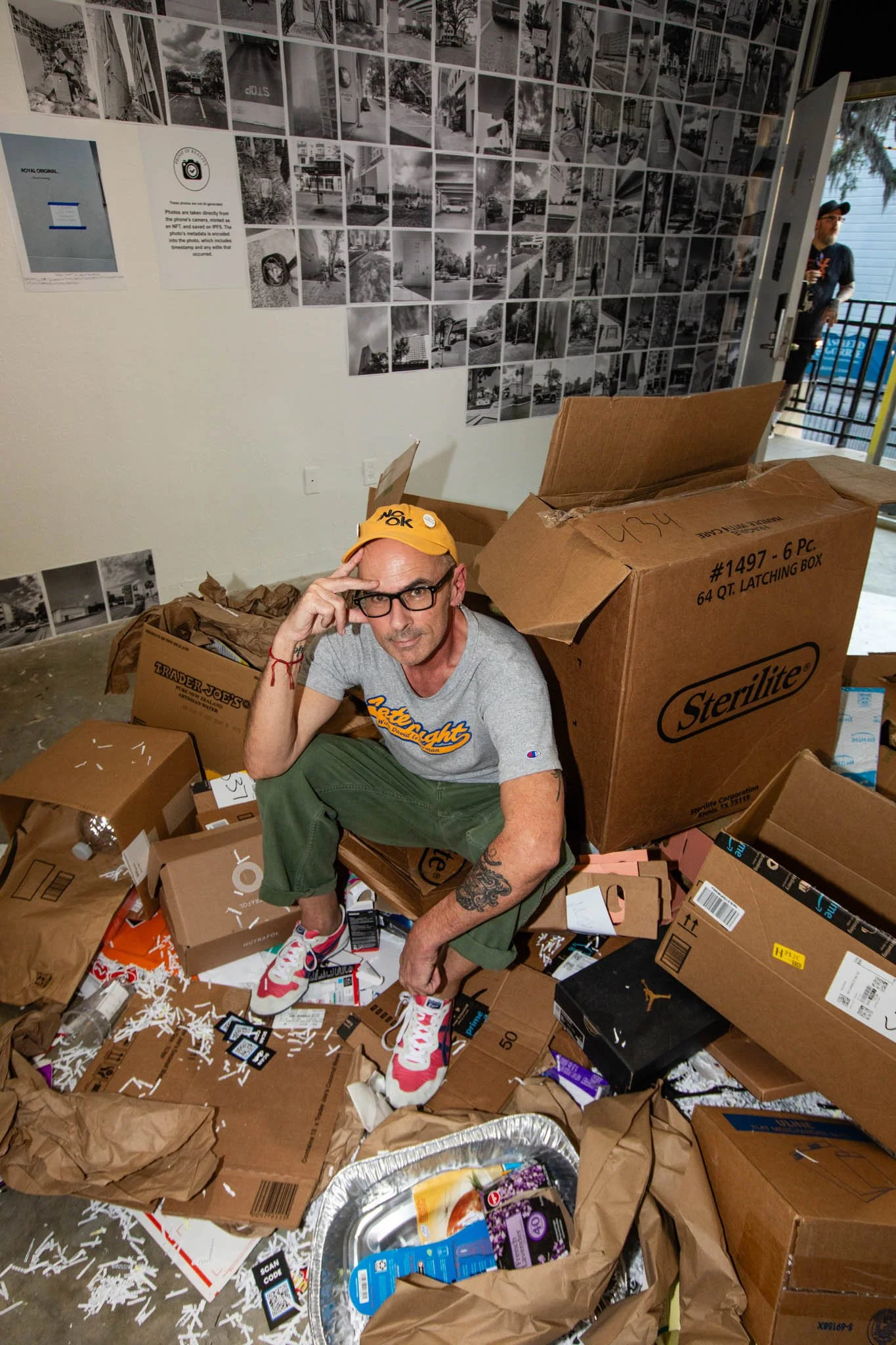 A man sits amidst a large pile of cardboard boxes and packaging paper, wearing glasses, a yellow cap, a light gray t-shirt, and green pants, with a tattoo on his left arm. The background shows a wall decorated with numerous black and white photograph