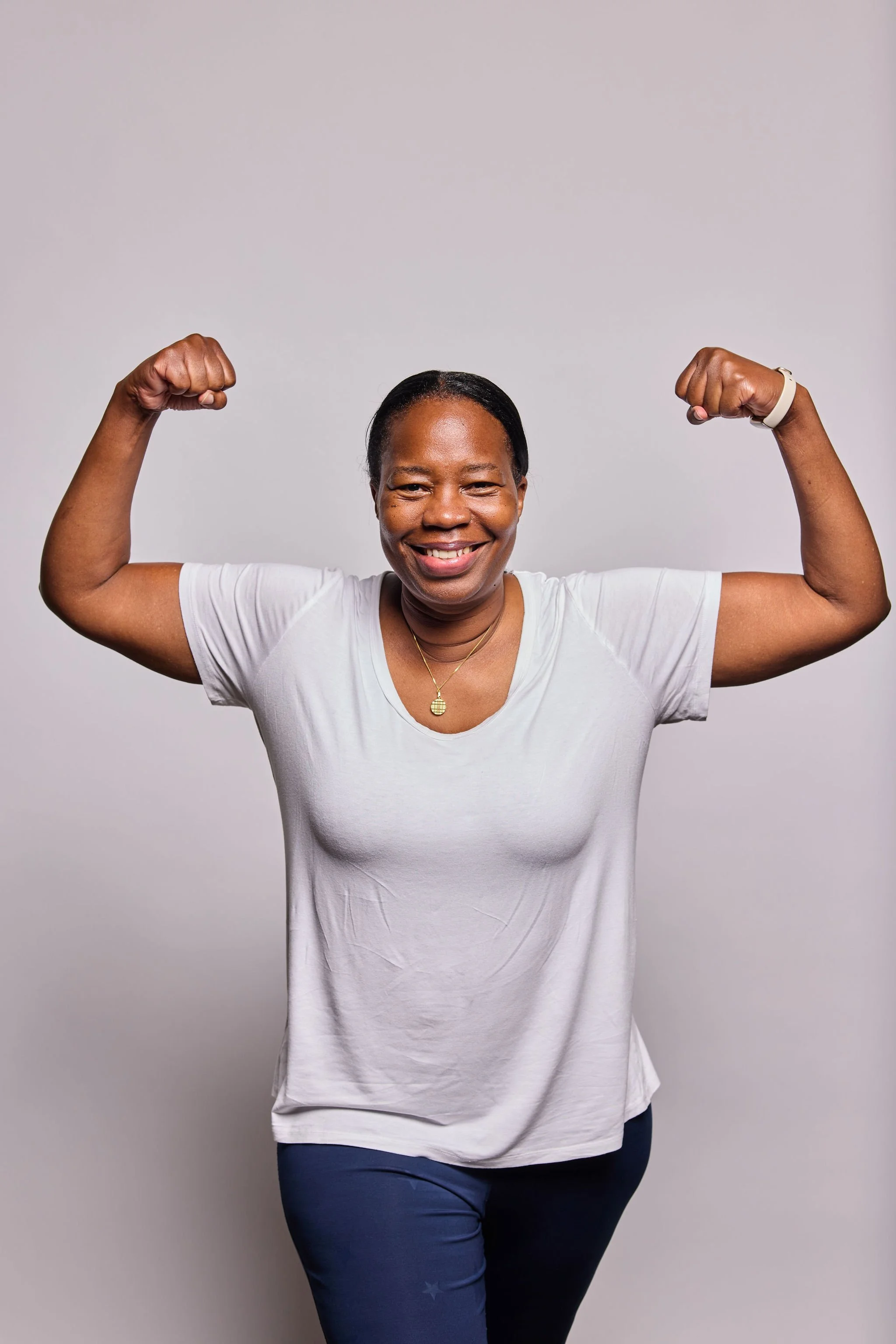 Woman smiling, flexing biceps, wearing a white t-shirt, dark blue pants, and a watch, standing against a plain background.