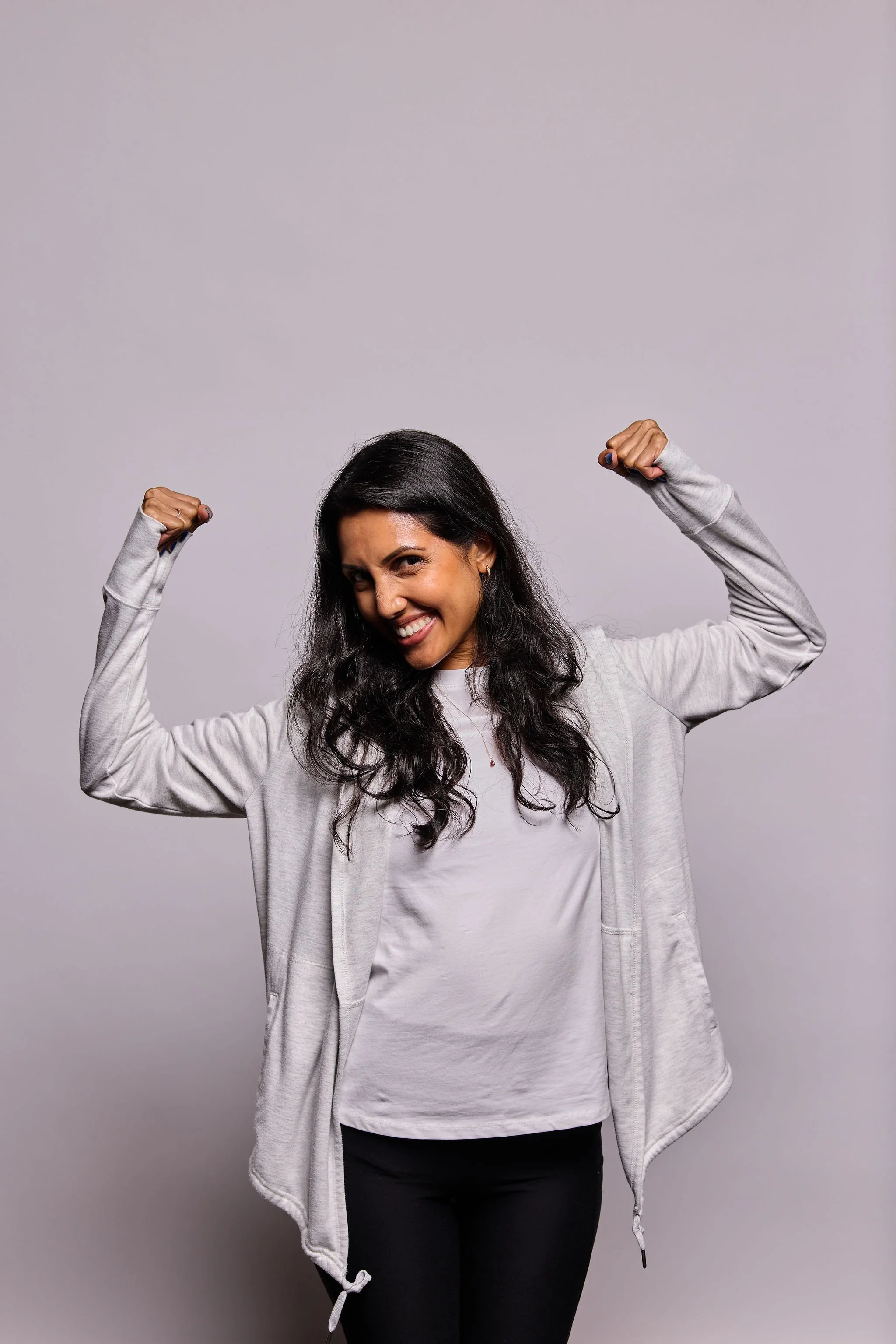 A woman with long dark hair smiling and flexing her biceps, wearing a light gray zip-up hoodie, light gray T-shirt, and black pants, standing against a plain light gray background.