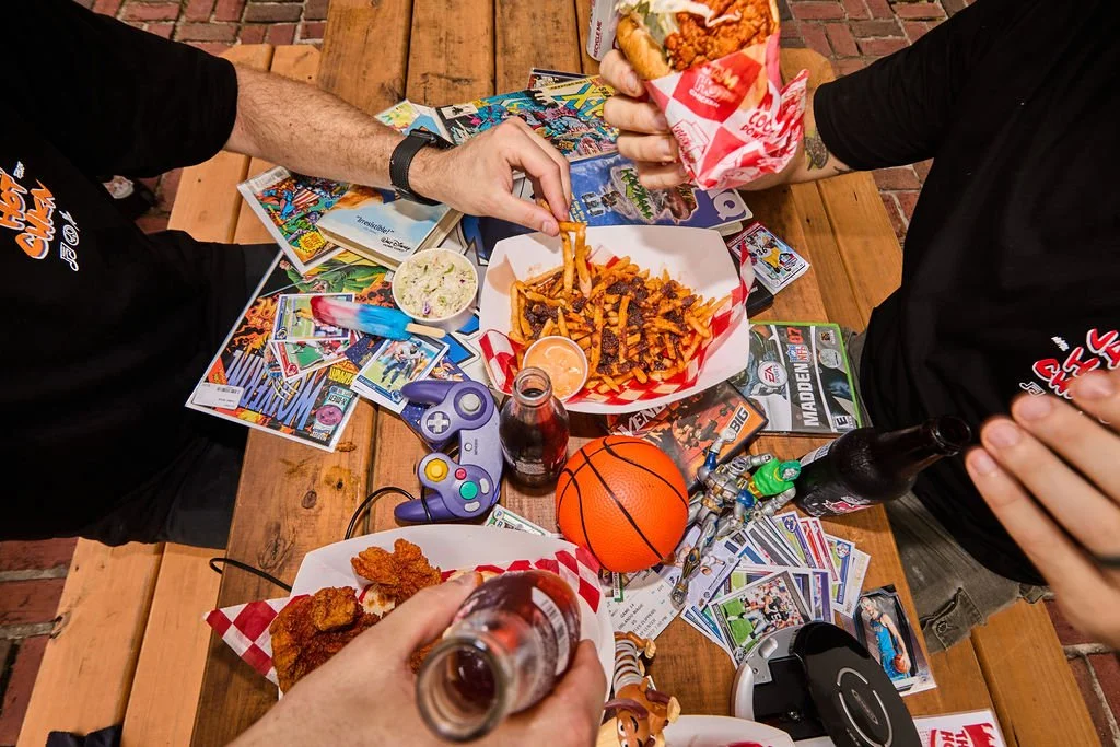 People having a meal with fried chicken, fries, and coleslaw on a wooden table decorated with trading cards, a basketball, a game controller, and soda bottles, with some hands holding drinks and food.
