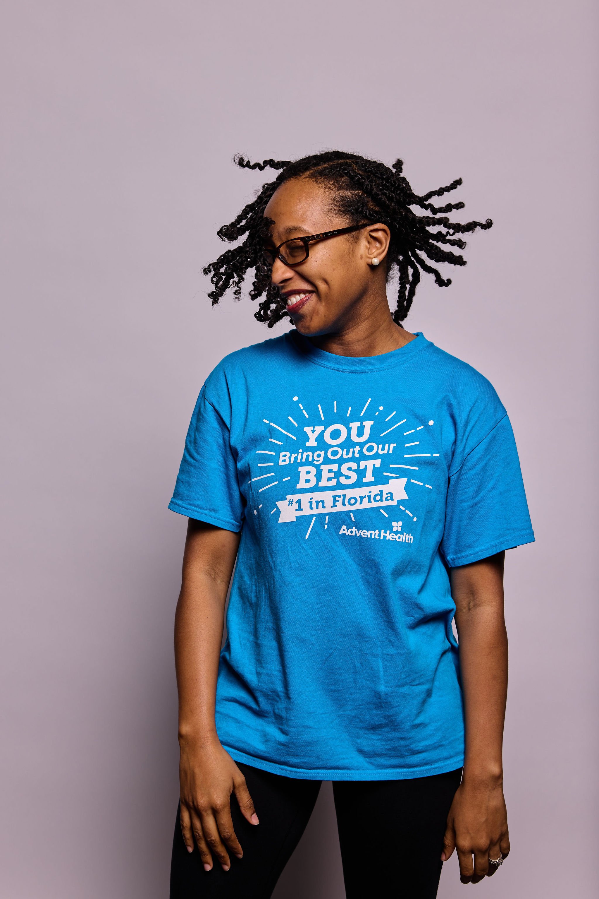 A woman with glasses smiling, wearing a blue T-shirt that says 'You Bring Out Our Best' and '1 in Florida' with a logo, standing against a plain gray background.