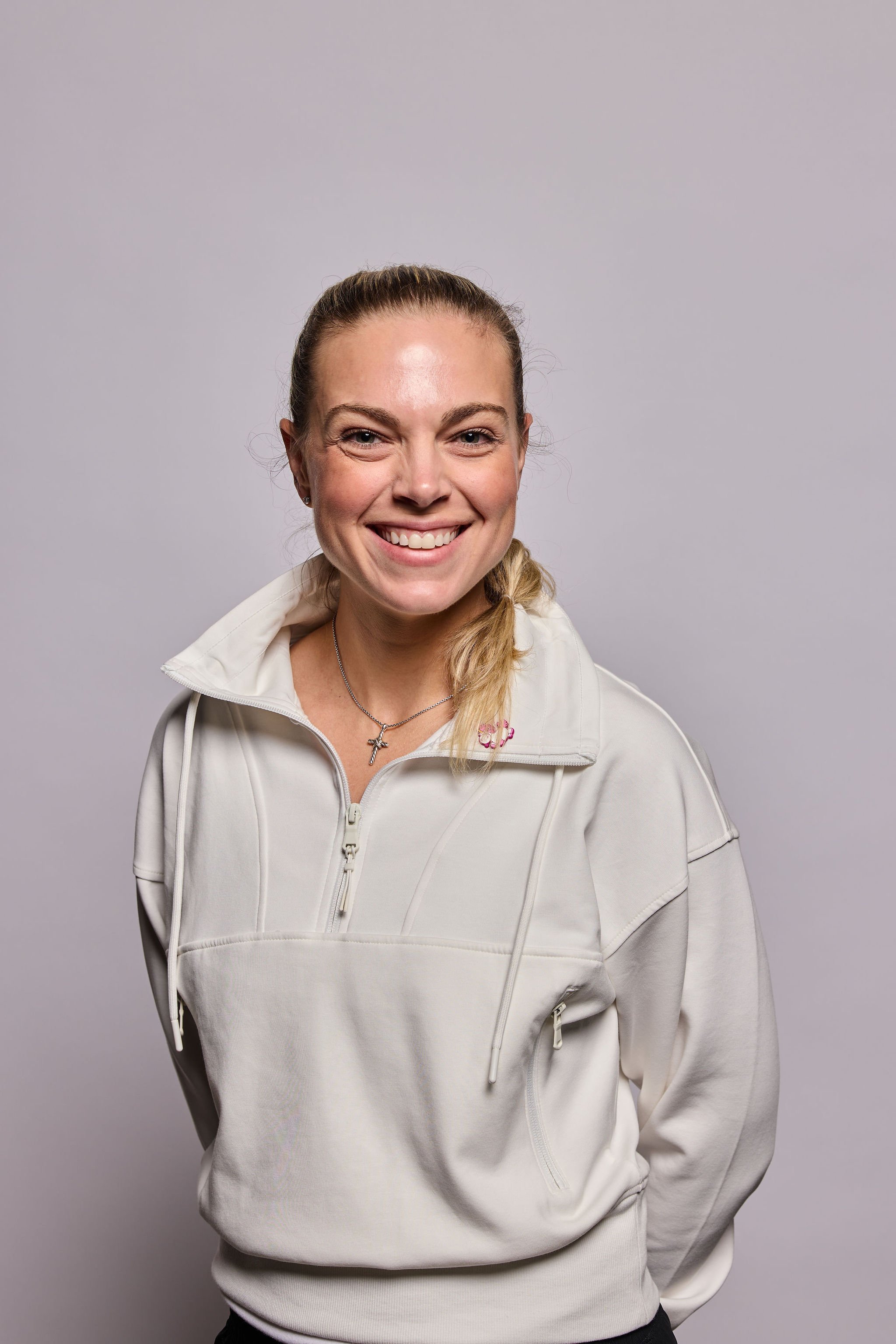A woman smiling, wearing a white zip-up hoodie and a silver necklace, against a plain gray background.