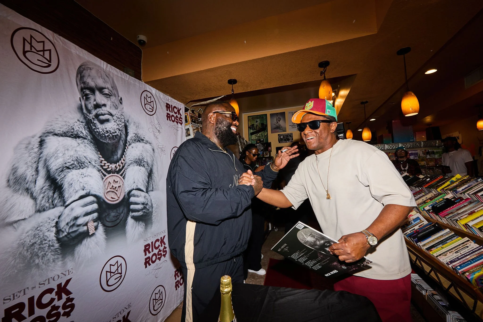 Two men shaking hands at a book signing event in a bookstore, with a large poster of Rick Ross in the background.