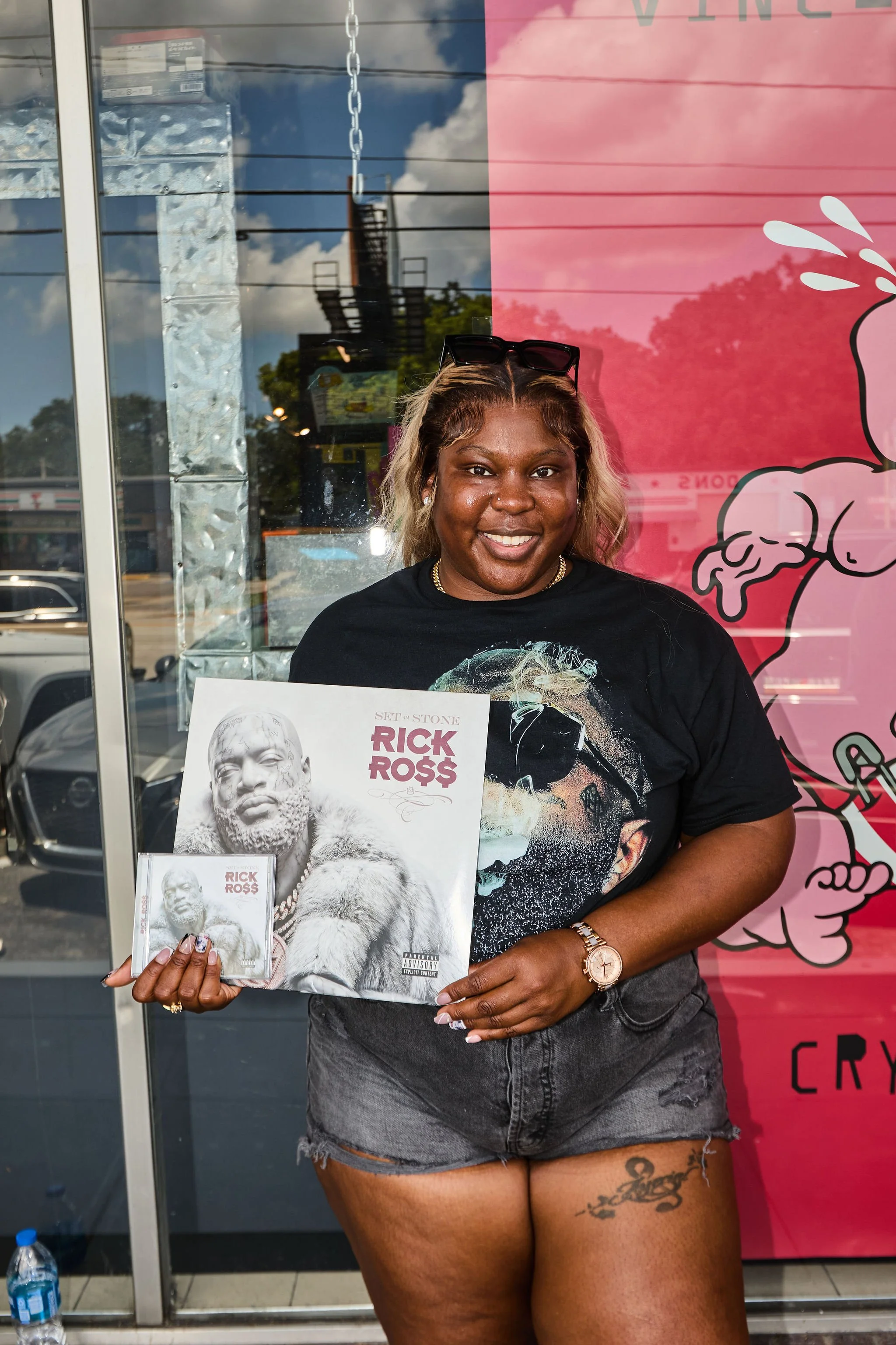 A woman with blonde hair, sunglasses on her head, wearing a black graphic T-shirt and denim shorts, holding a music album titled "Set in Stone" by Rick Ross, standing in front of a store window with pink and black artwork and a reflection of the sky and trees.