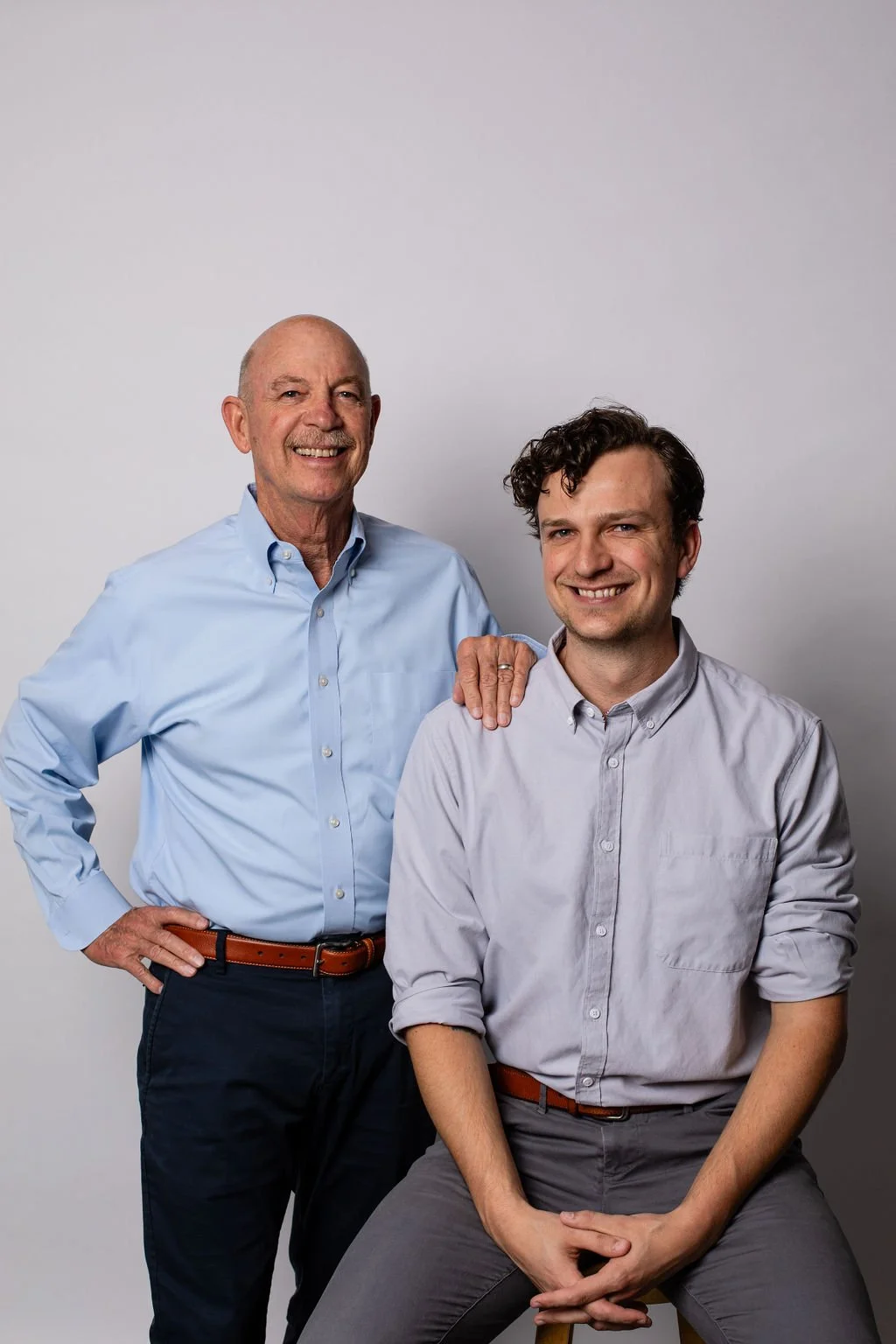 Two men smiling, one older and one younger, posing against a neutral background, with the older man placing his hand on the younger man's shoulder.