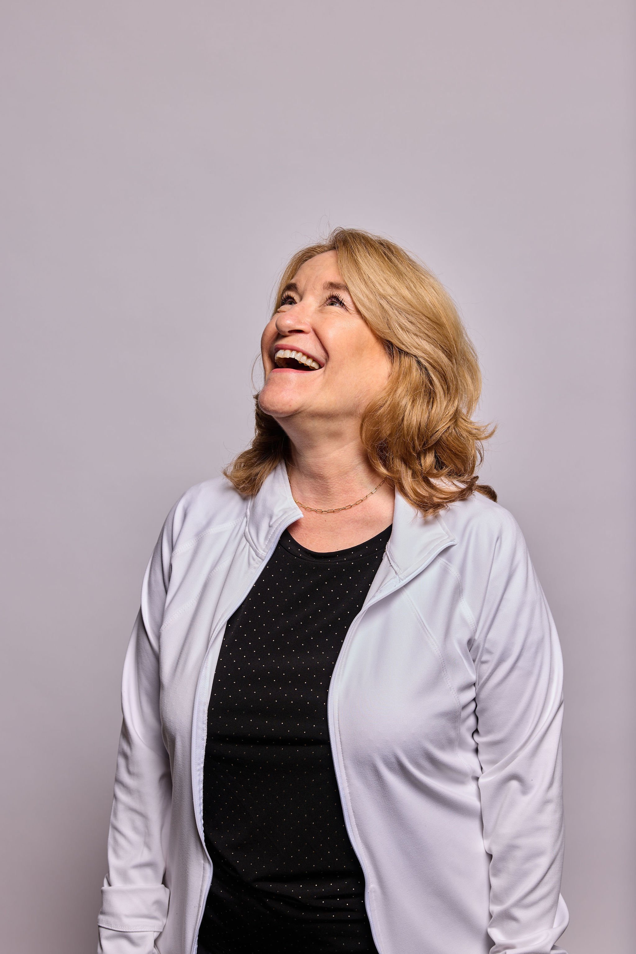 A woman with shoulder-length light brown hair smiling and looking upward, wearing a white jacket over a black shirt with small white dots, against a plain light gray background.