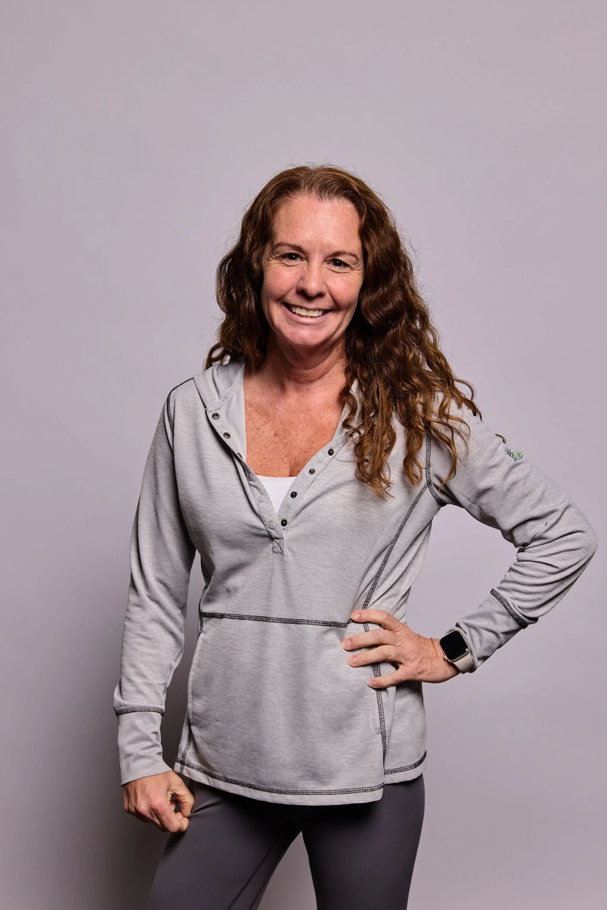 A smiling woman with long, curly reddish-brown hair wearing a light gray athletic jacket with black accents, gray leggings, and a smartwatch, standing against a plain gray background.