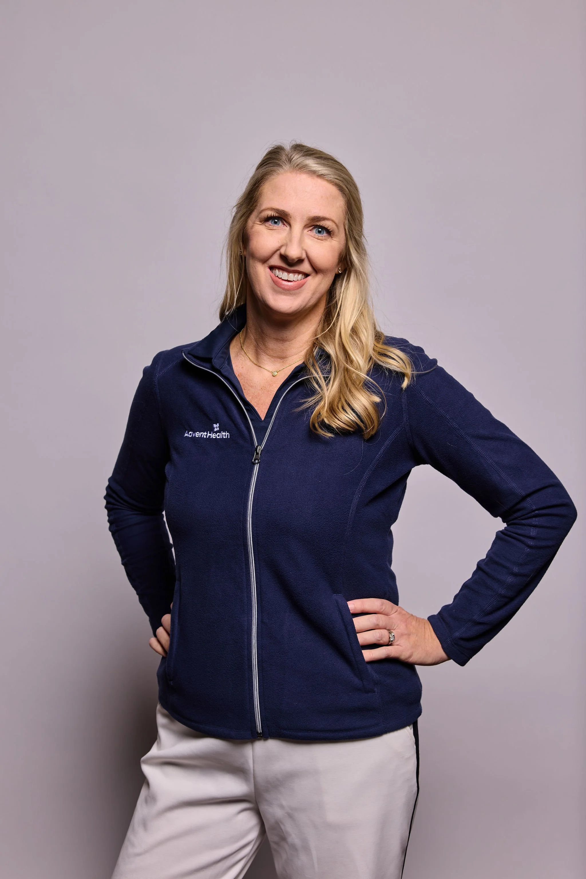 Woman with blonde hair wearing a navy blue jacket and white pants, standing against a gray background.