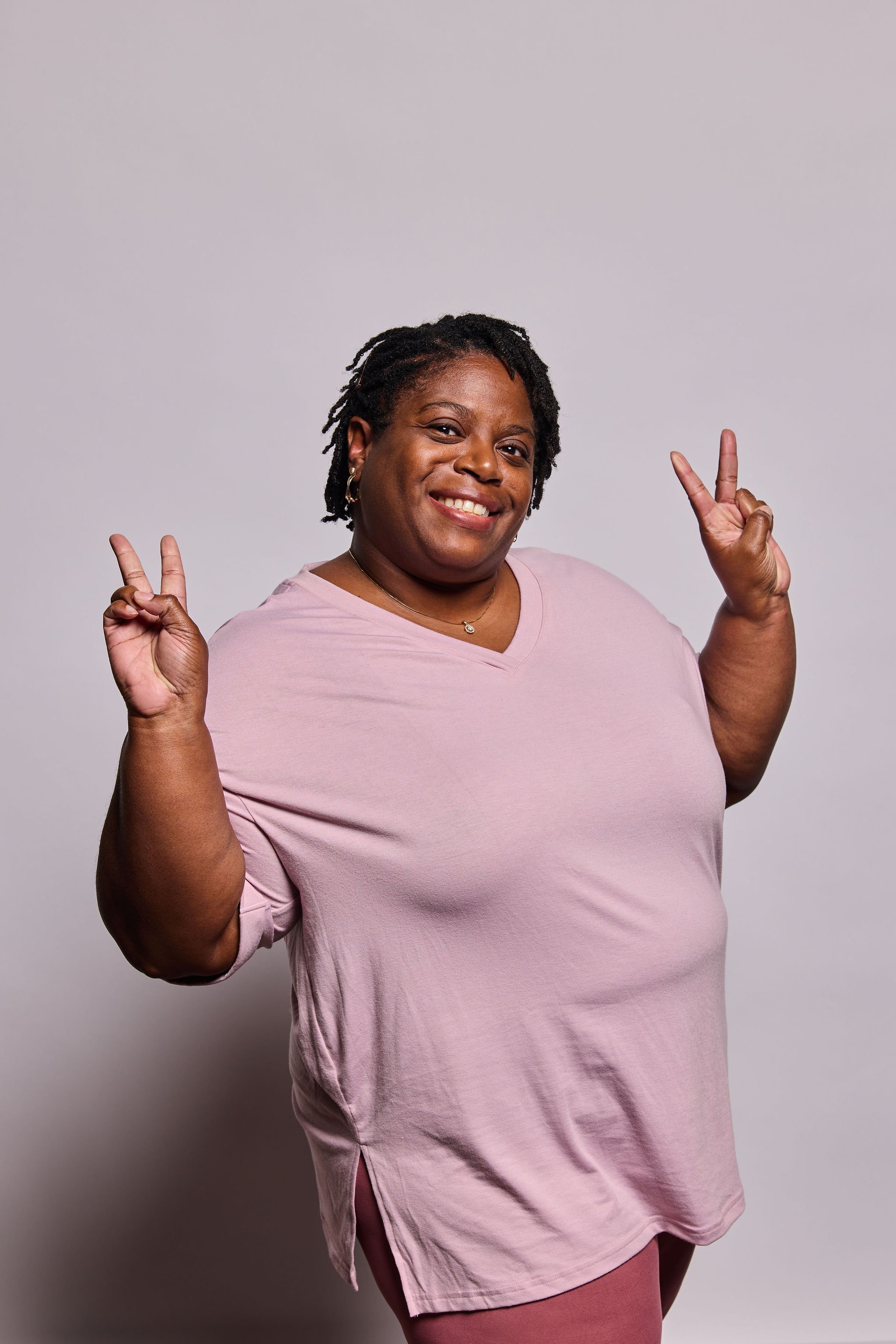Smiling woman with short dreadlocks wearing a light pink t-shirt making peace signs with both hands against a neutral background.