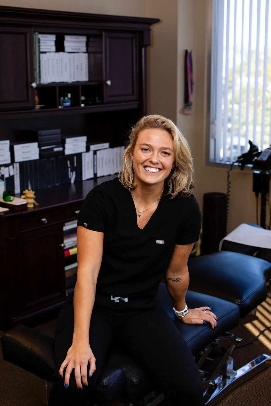 A smiling woman with blonde hair, dressed in black scrubs, sitting on an examination table in an office or clinic and looking at the camera. Behind her are shelves with files and small medical supplies, a window with vertical blinds, and medical equipment.