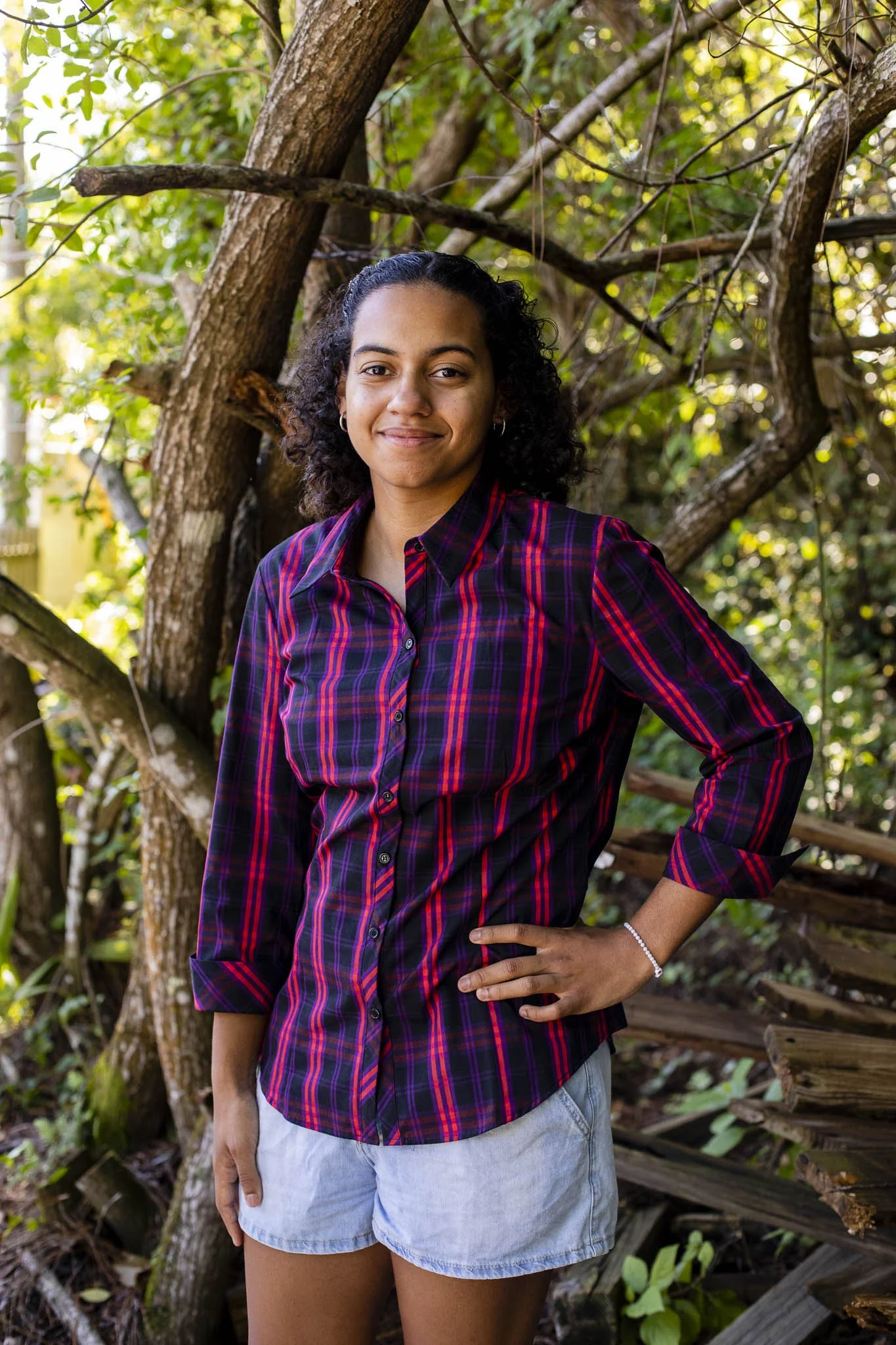 A woman with curly dark hair wearing a red and black plaid shirt and light denim shorts, standing outdoors among trees and branches, smiling with her right hand on her hip.