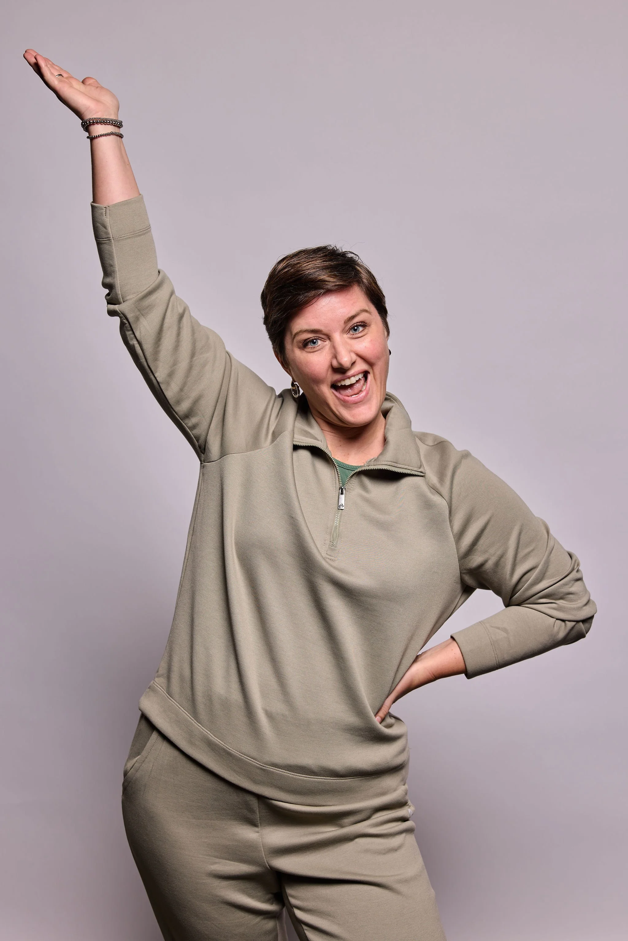 A woman with short brown hair wearing a beige tracksuit, smiling and striking a confident pose with one arm raised and on her hip, against a plain gray background.