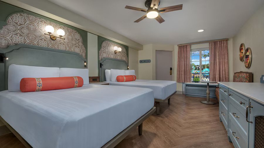 New room as of 2026 at Port Orleans French Quarter. White bed linens, orange pillows, and hardwood floors.