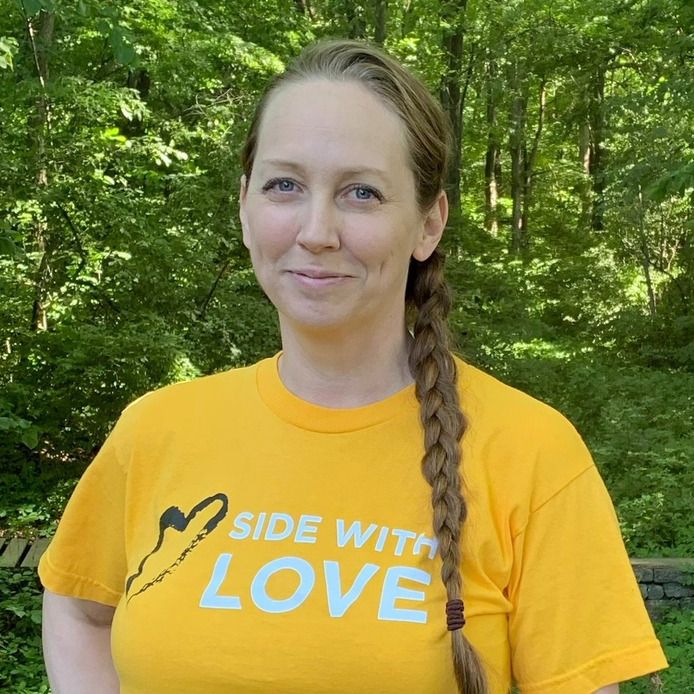 a person in a yellow Side With Love shirt stands in front of green trees and bushes. They have hair in a braid and are smiling.