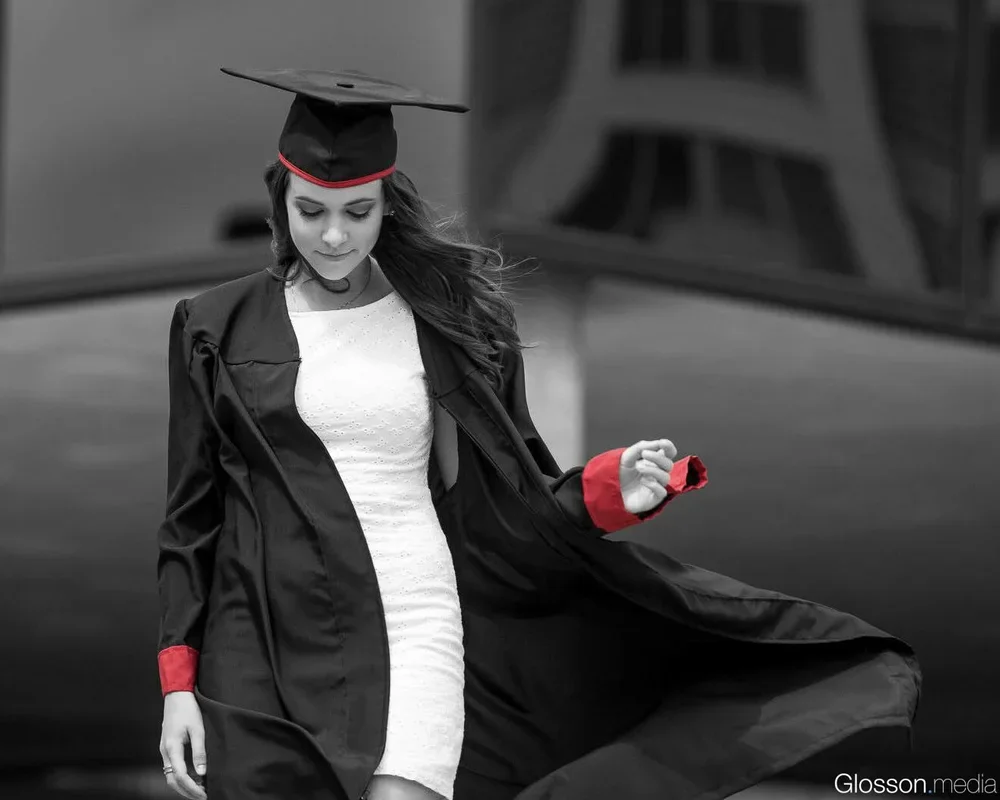 A young woman in a graduation cap and gown, holding the gown with one hand, looking down, with a modern building in the background.
