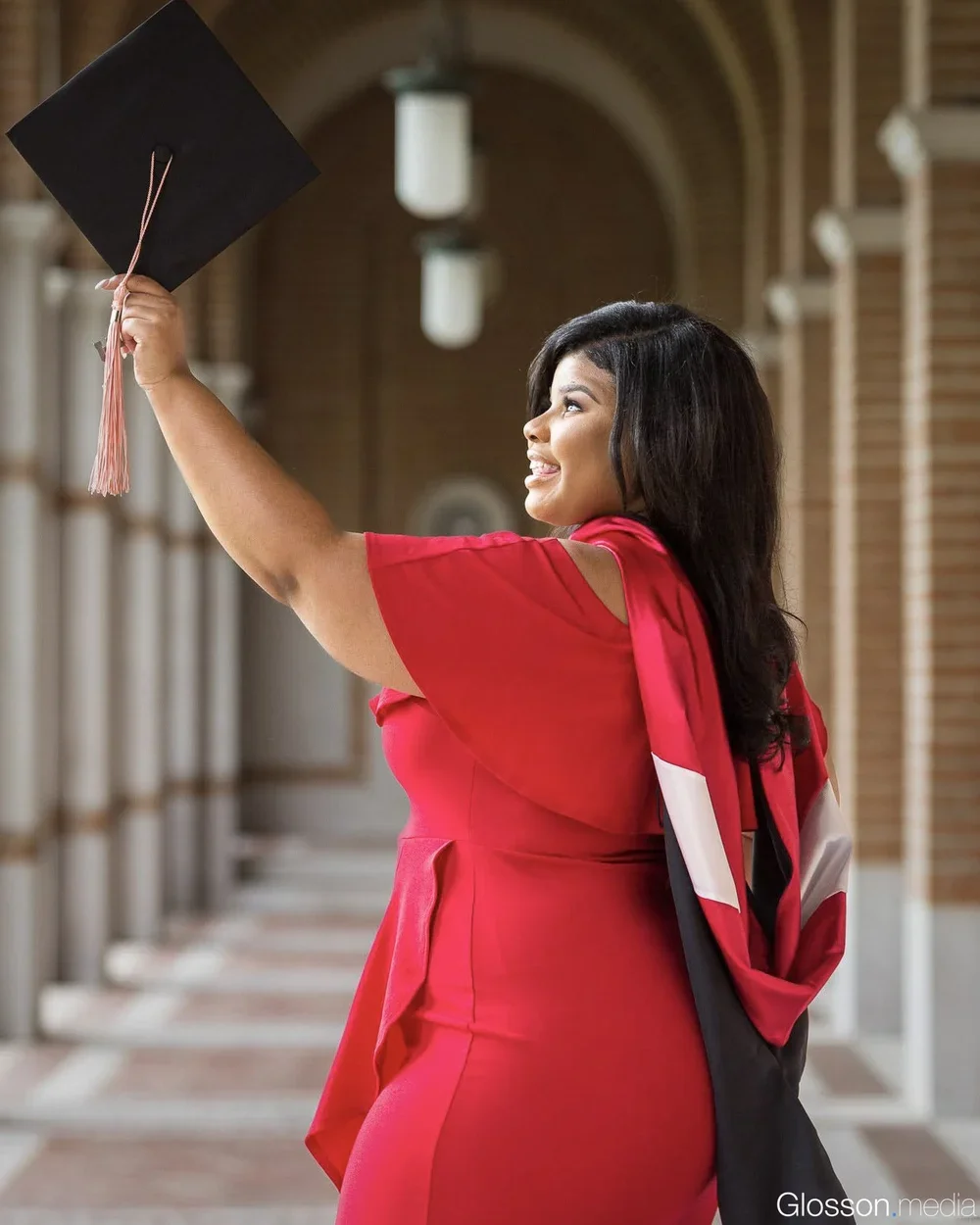 A woman in a red dress celebrating her graduation, holding a graduation cap in an indoor corridor with brick walls and arched ceilings.
