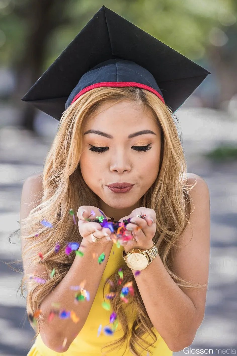 A young woman wearing a graduation cap and yellow dress blows colorful confetti from her hands, celebrating her graduation.