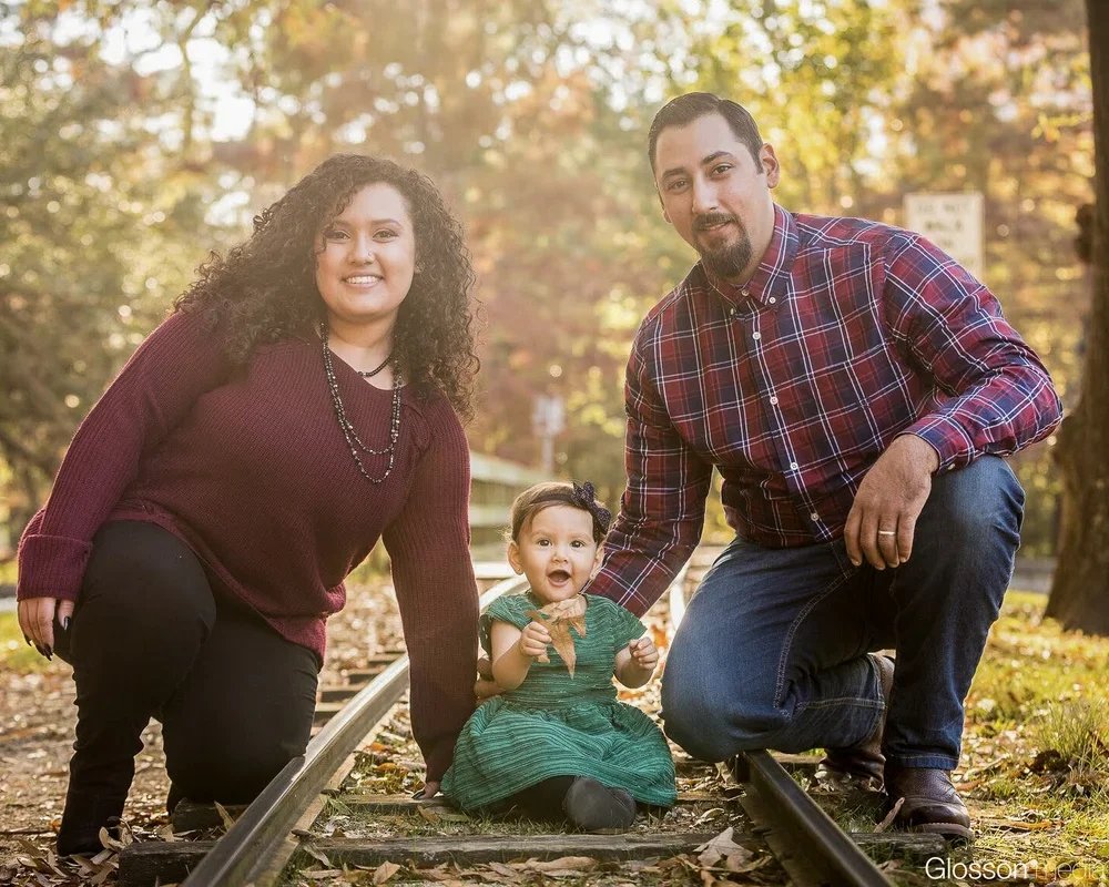 Family of three posing on railroad tracks in autumn. The woman has curly brown hair and wears a maroon sweater, the man has dark hair and wears a red plaid shirt, and the toddler girl has a black bow in her hair and wears a green dress, all smiling o