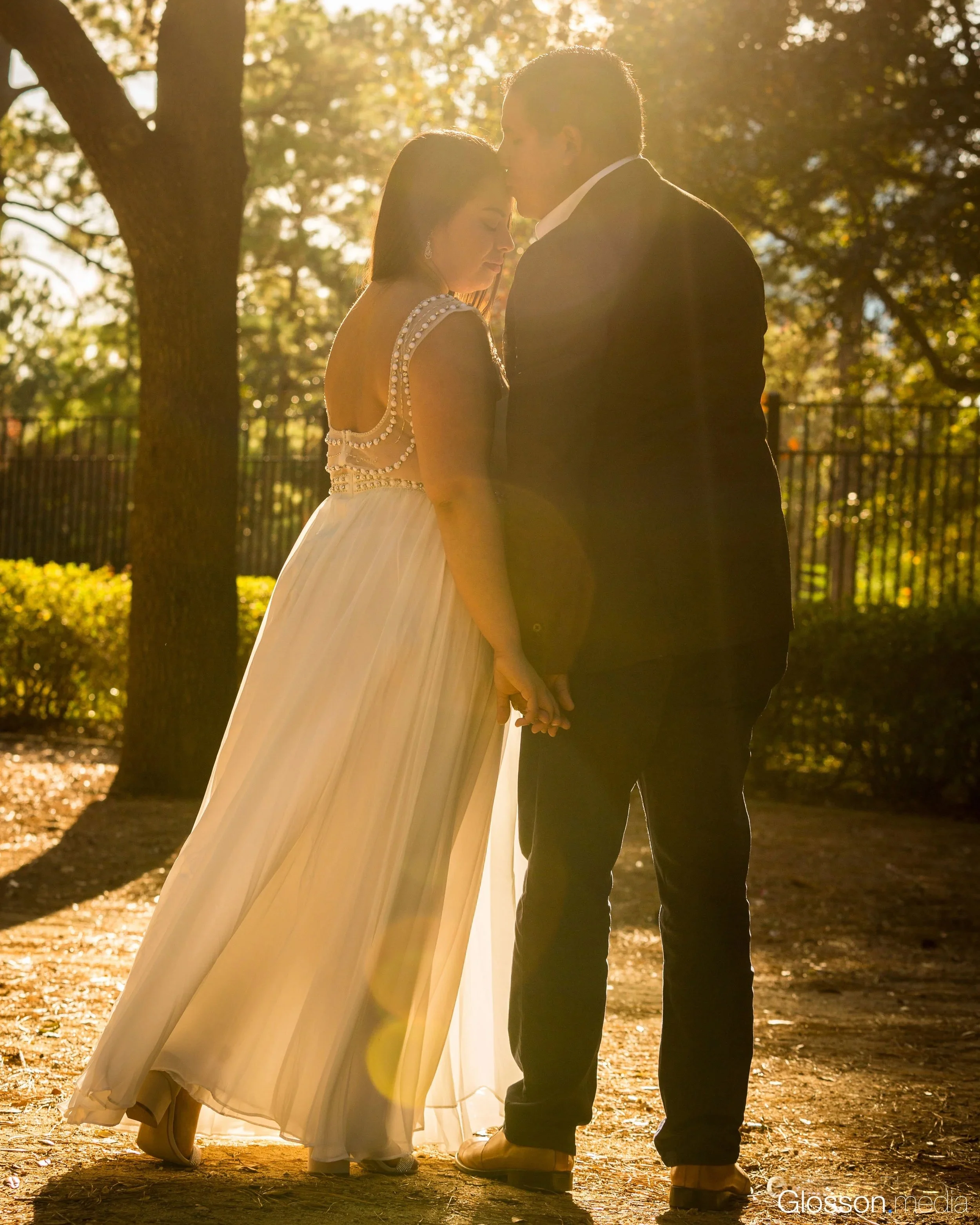 A couple dressed in wedding attire holding hands and standing close, with their foreheads touching in a park during sunset.