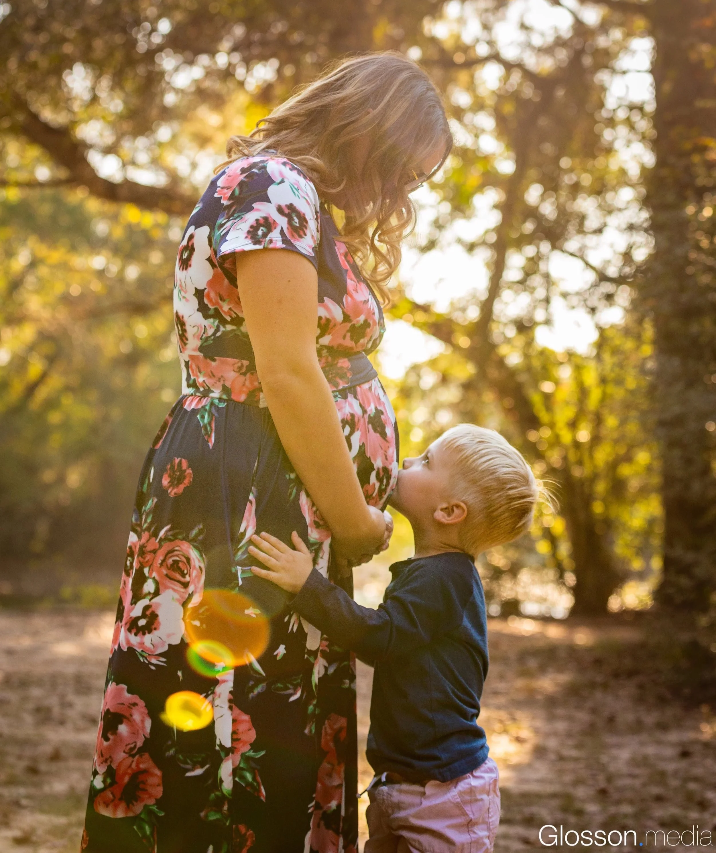 A woman with long, curly hair wearing a floral dress stands outdoors in a sunlit park, pregnant and gently holding her belly. A young boy with blond hair wearing a dark jacket and pink pants kisses her belly. The background shows trees with sunlight 