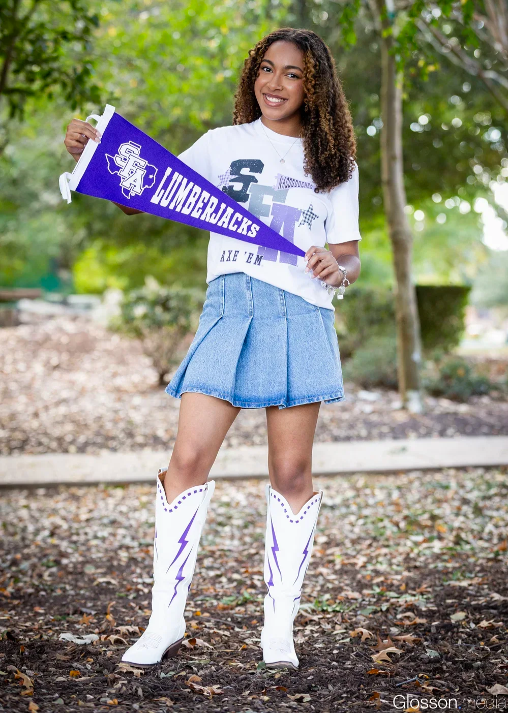 Young woman smiling outdoors holding a purple JUMBERJACKS pennant, wearing a white T-shirt, blue denim skirt, and white cowboy boots with purple lightning bolts, trees and foliage in the background.