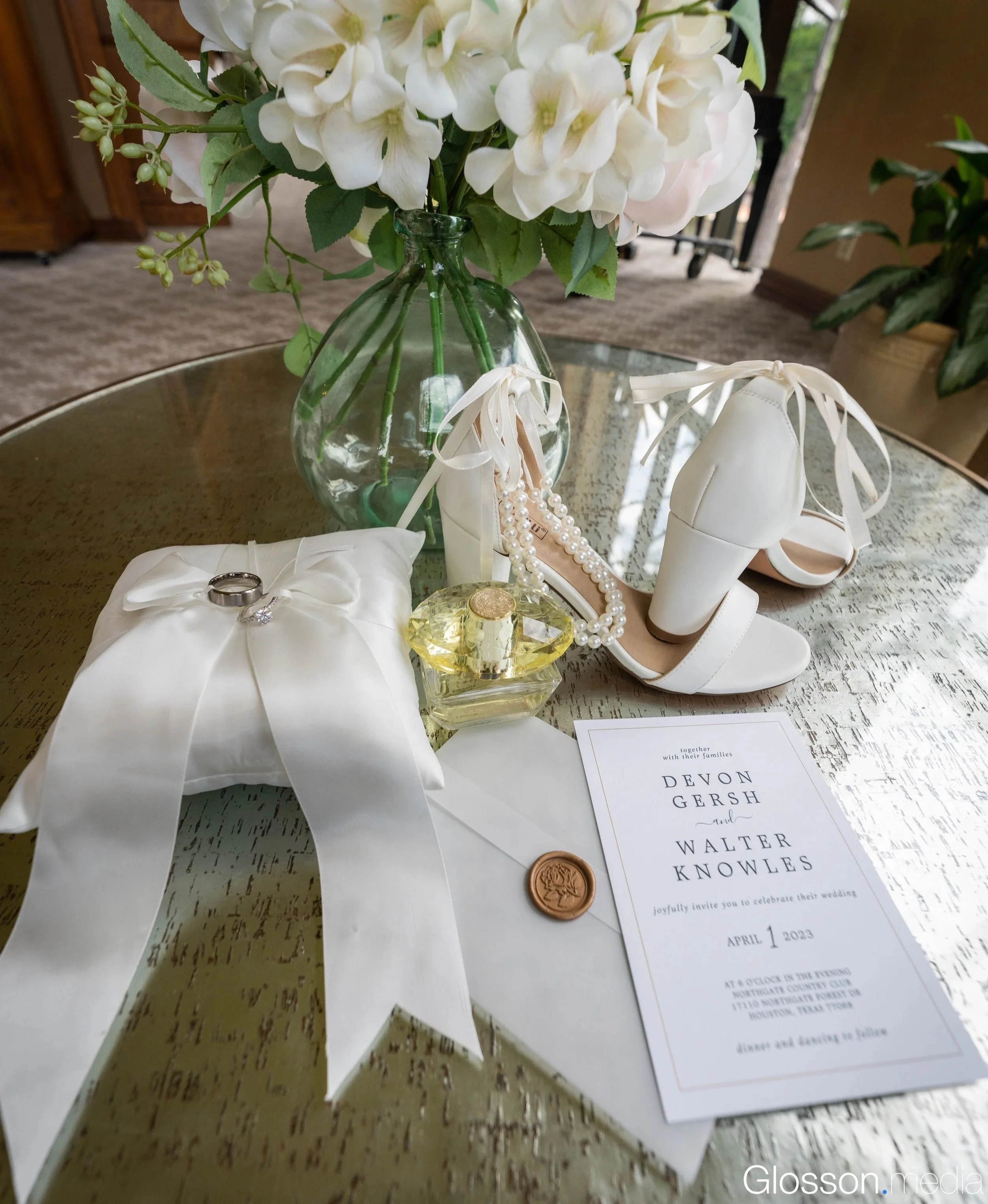 Wedding invitation, white high-heeled shoes with ankle ribbons, pearl necklace, perfume bottle, wedding rings on satin pillow, vase of white flowers, and a wax seal on a gold table.