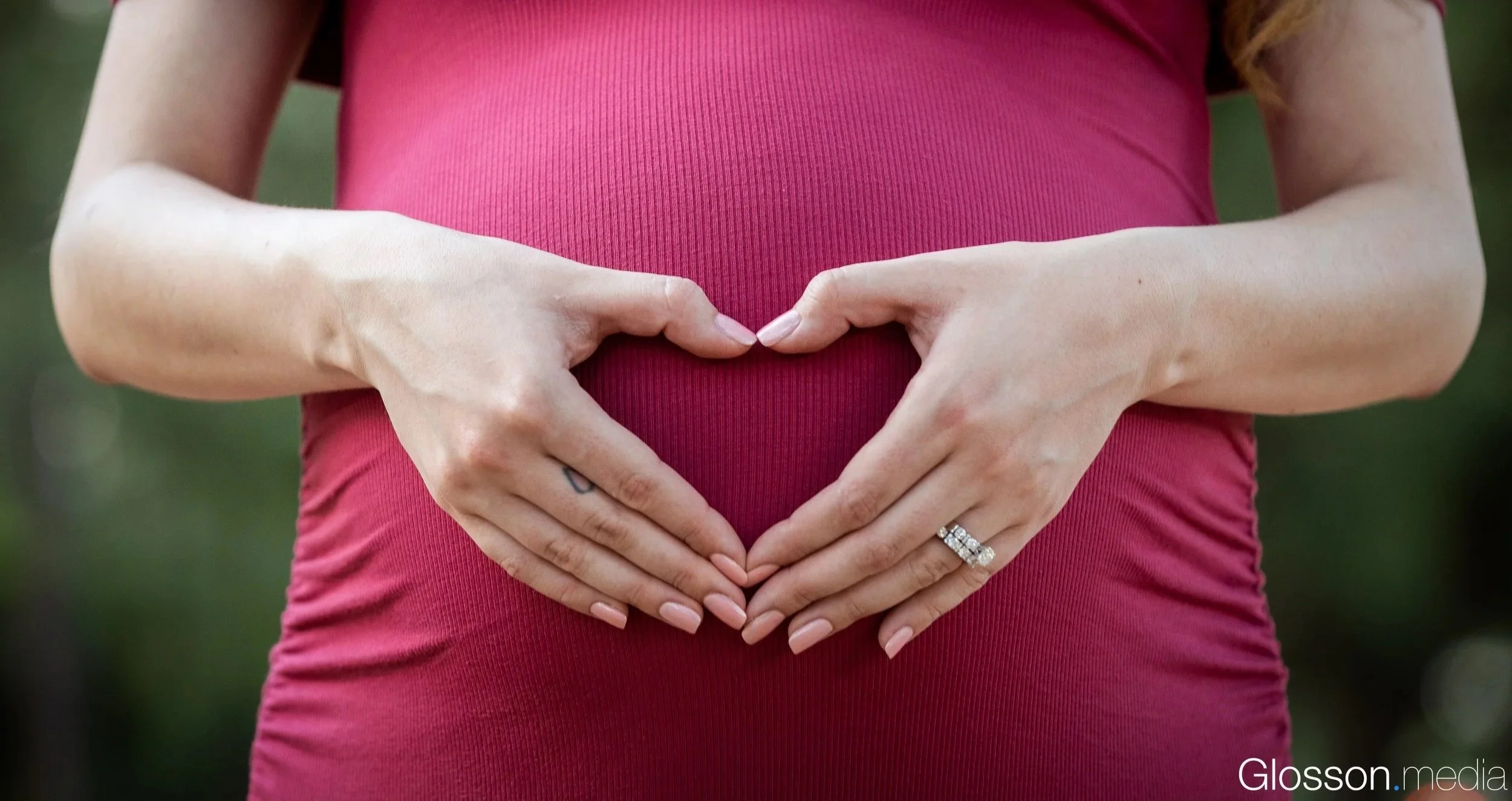 A pregnant woman in a magenta dress forms a heart shape with her hands on her belly.