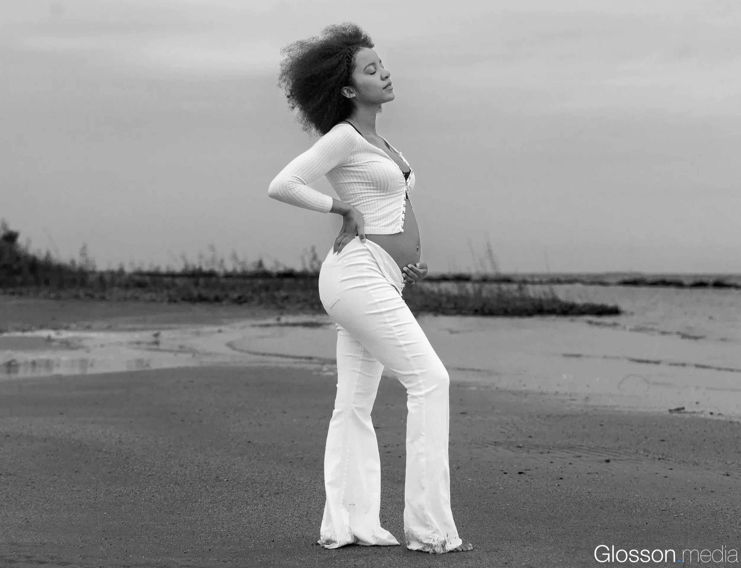 A woman stands on a beach with her eyes closed, wearing white pants and a long-sleeve top, with her left hand on her waist and her right hand resting on her stomach, facing slightly to the right. The landscape includes sand, water, and distant indust
