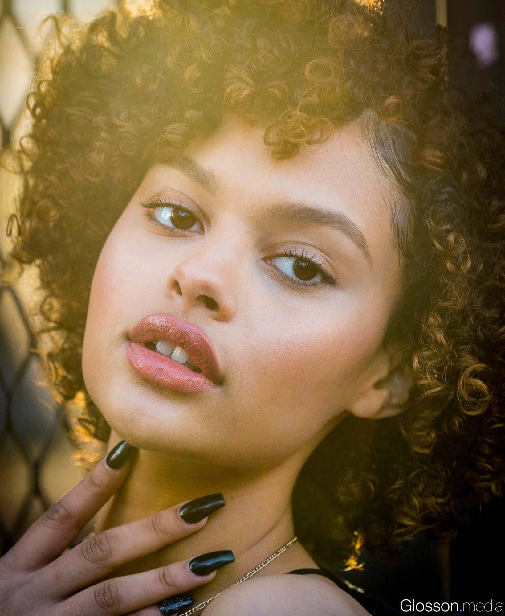 Close-up portrait of a woman with curly hair, makeup, and black nails, posing sensually with her hand near her neck.
