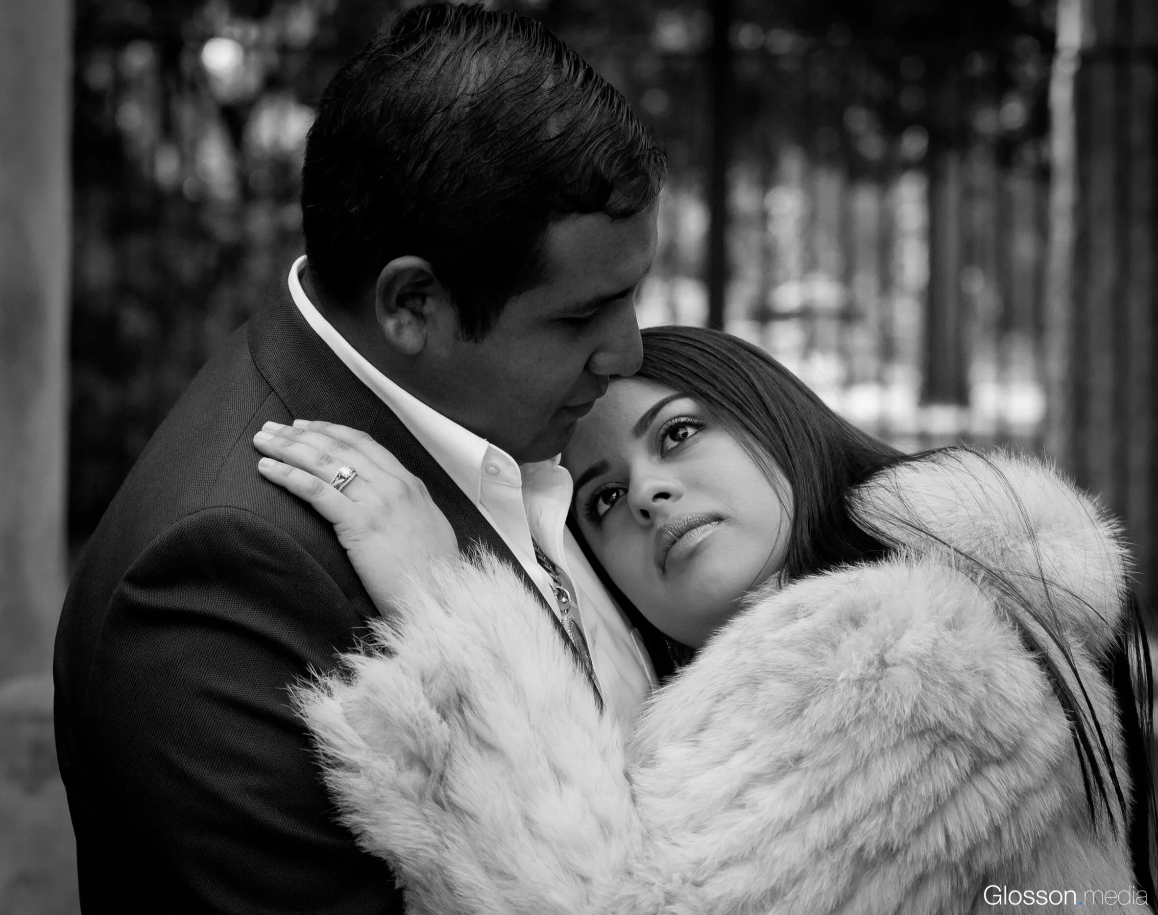 A man and woman embrace outdoors, with the woman gazing into the man's face. The woman is wearing a fur coat, and the man is dressed in a suit.