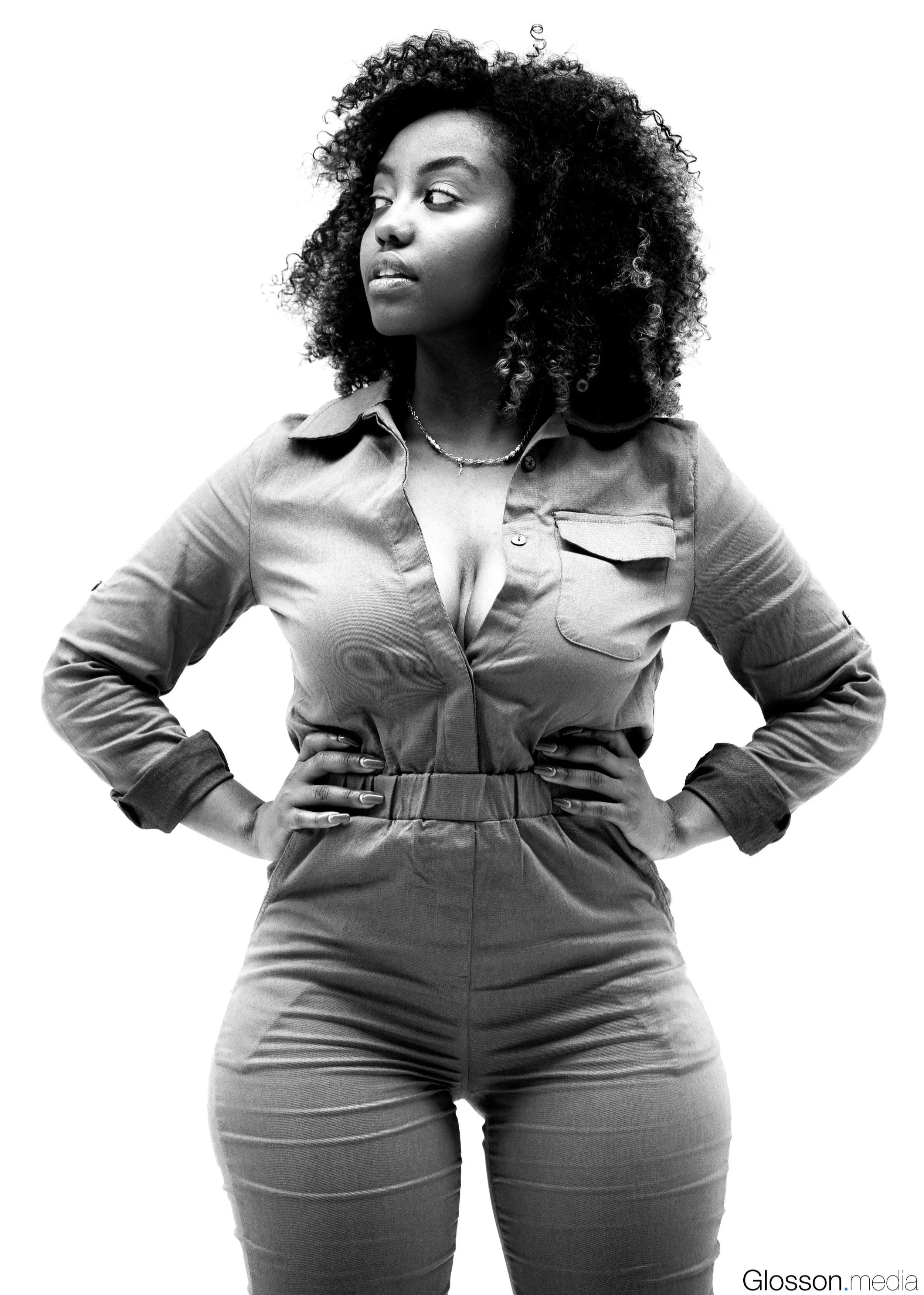 A confident woman with curly hair standing with her hands on her hips, wearing a long-sleeve jumpsuit, looking to the side, on a white background.