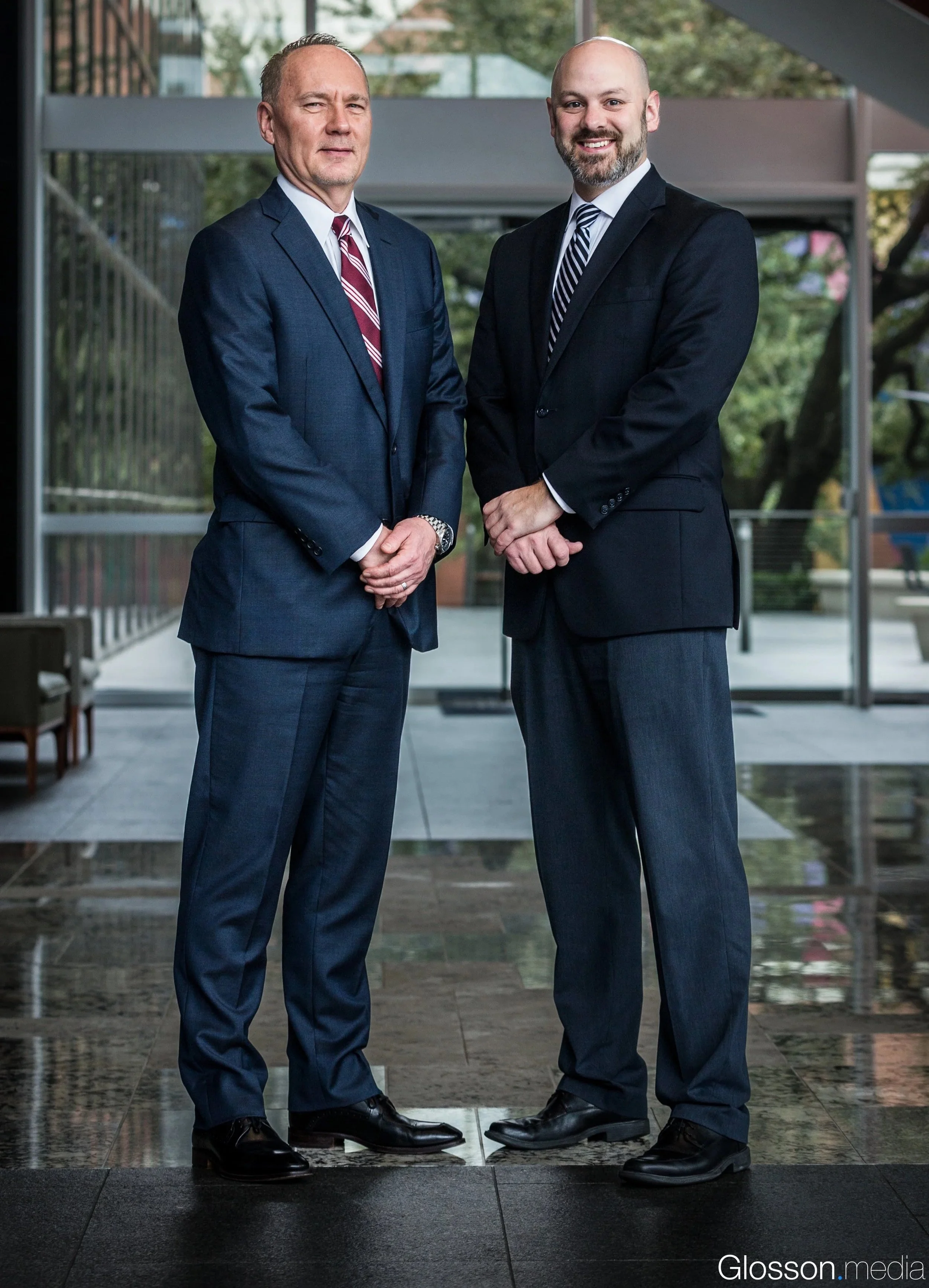 Two men in business suits standing outdoors, smiling, with trees and a building in the background.