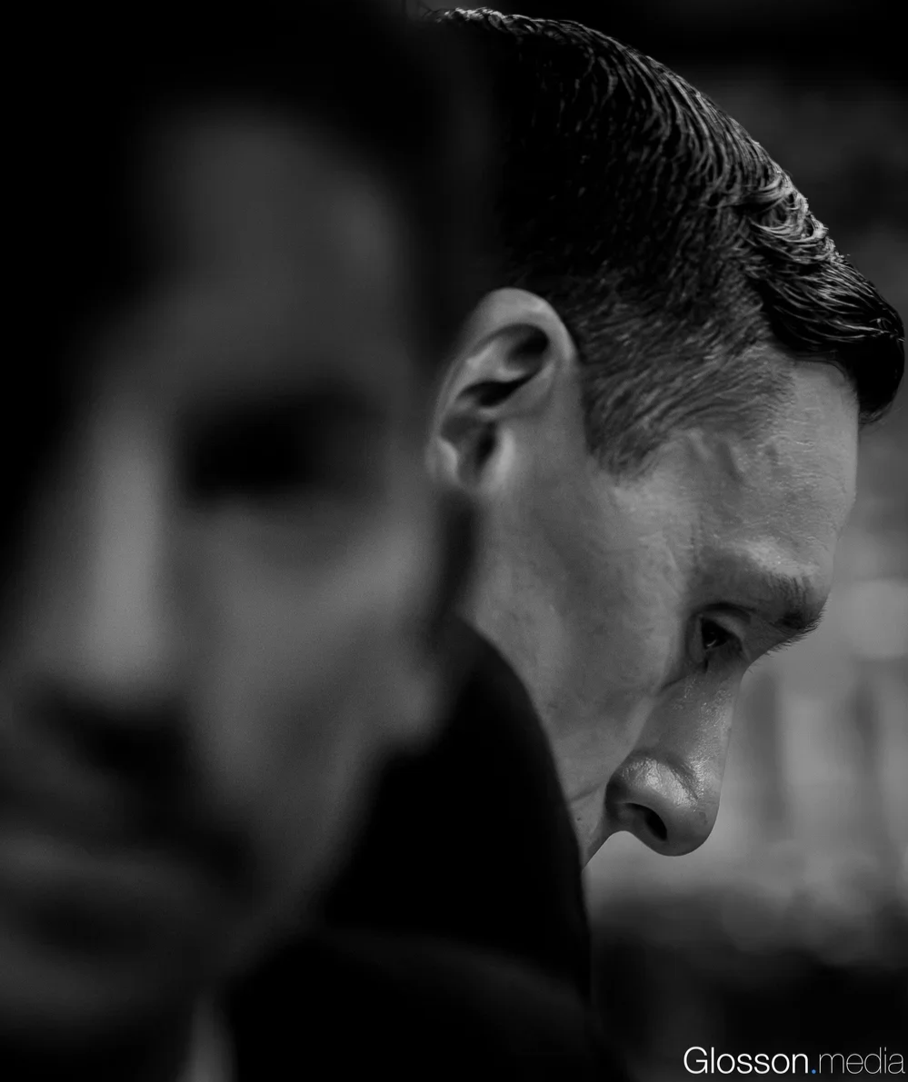 Close-up of two men's faces in black and white, with focus on the man in the background who has slicked-back hair and a serious expression.