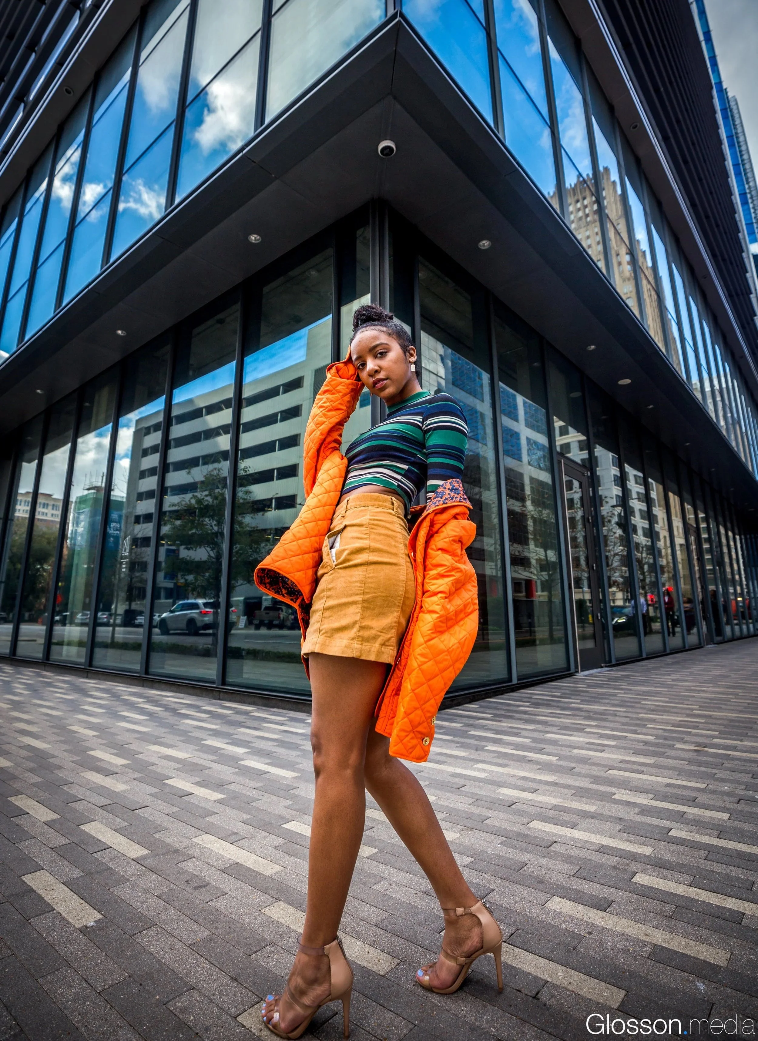 Young woman standing on city sidewalk in front of glass building, wearing striped crop top, a tan skirt, and high heels, with an orange jacket draped over her shoulder.