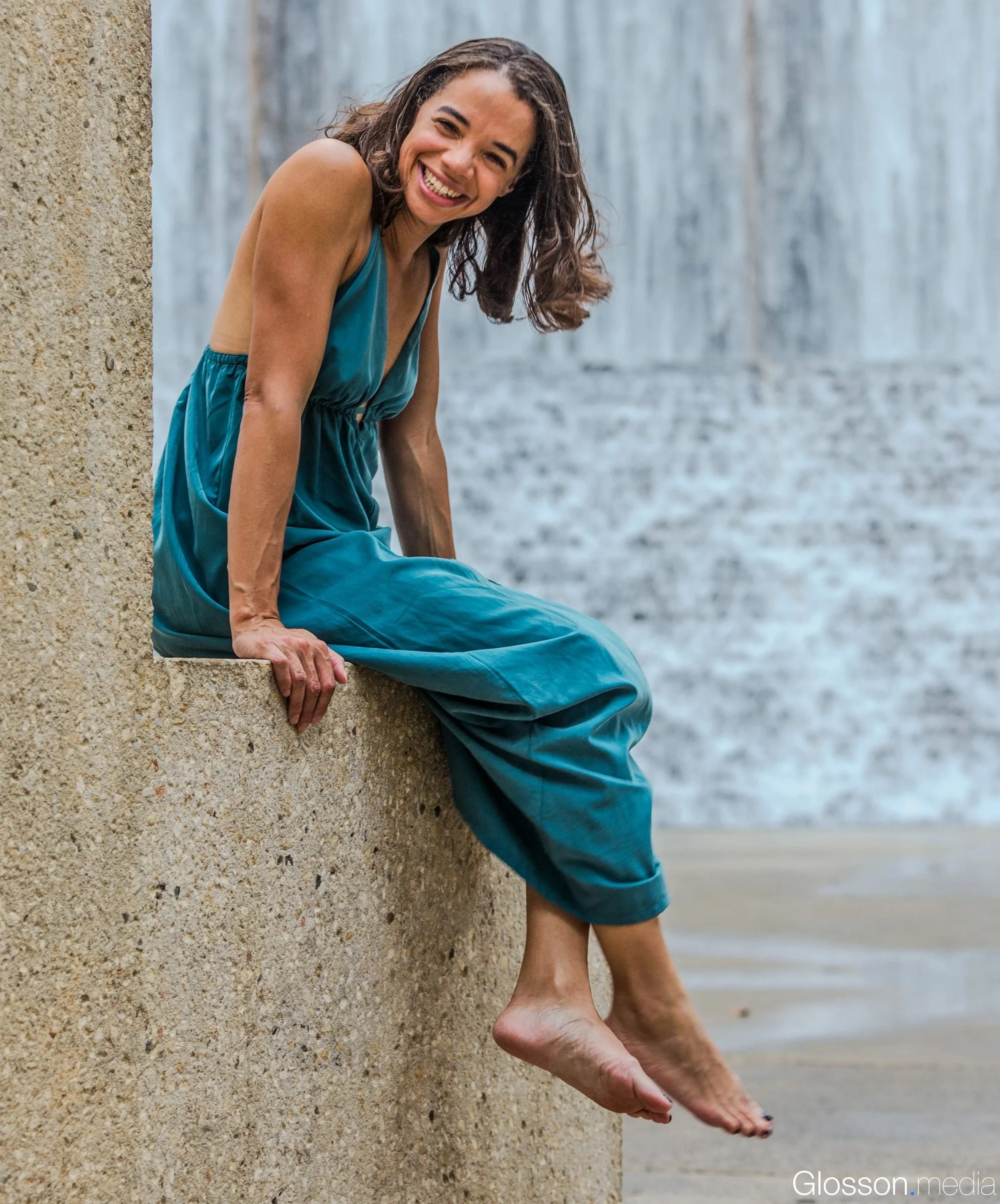 A woman with curly hair smiling, wearing a blue dress, sitting on a sandy beach next to a concrete structure, with water in the background.