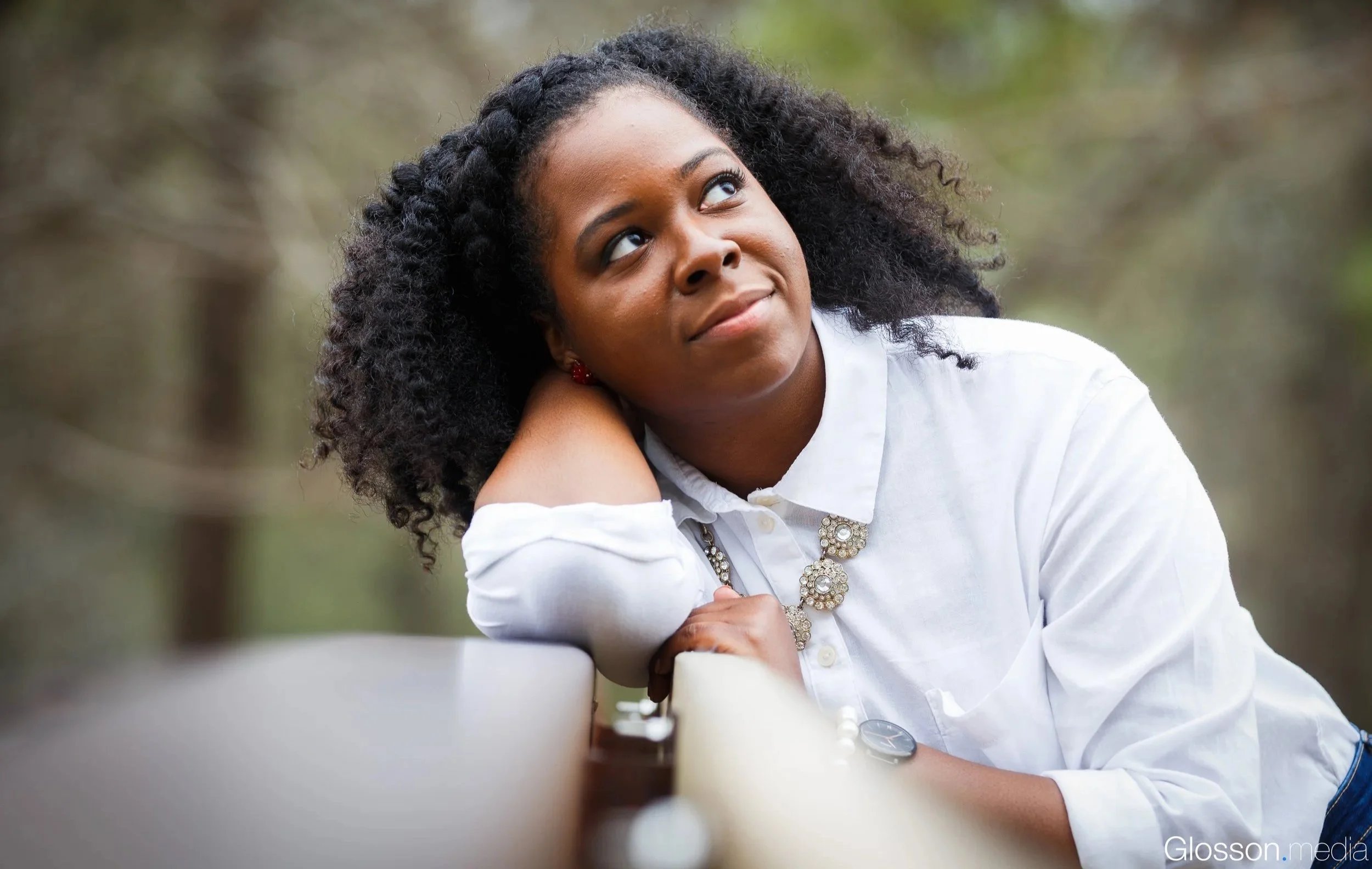 A woman with curly black hair and a white blouse with a decorative necklace leaning on a railing, looking to the side outdoors.