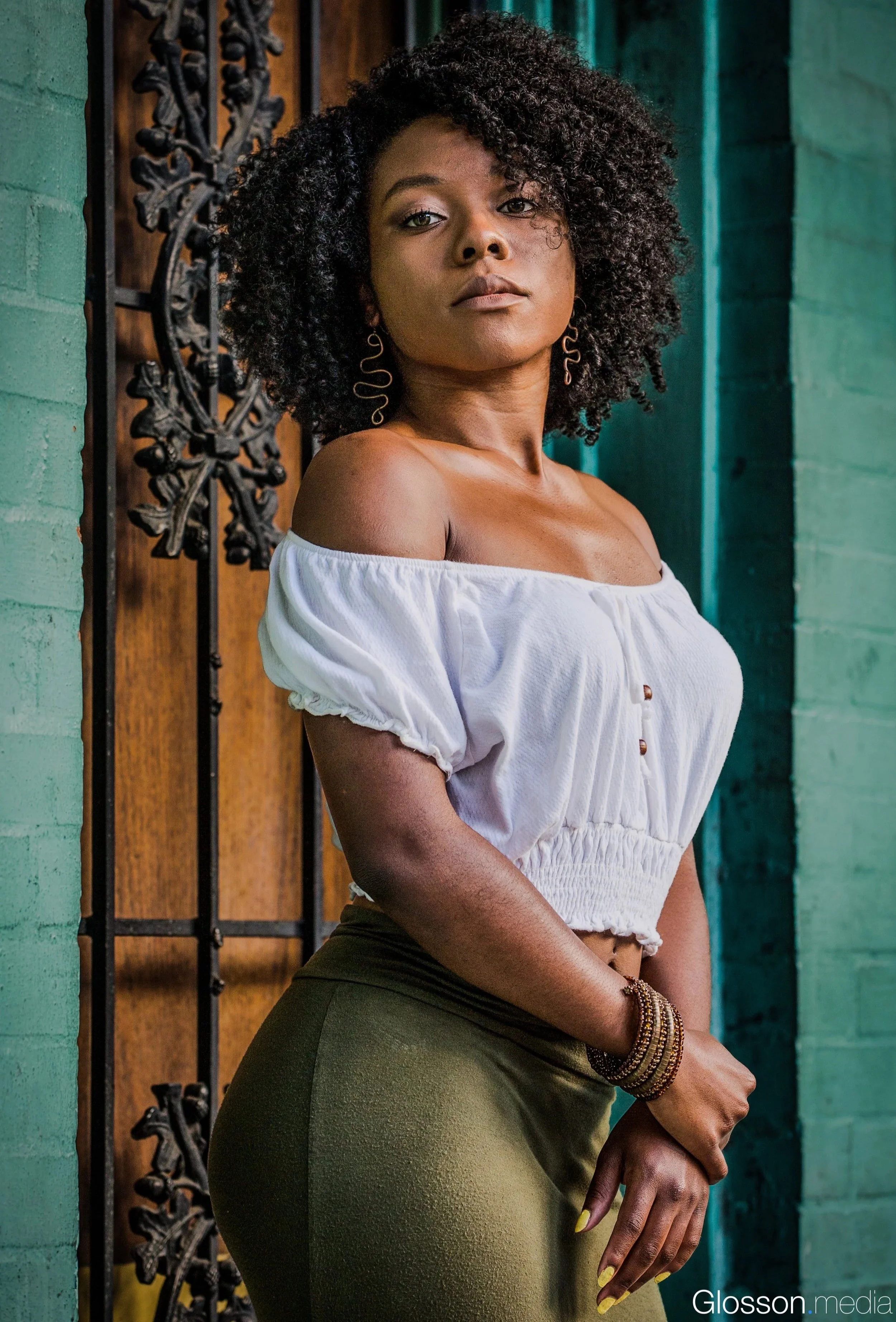 A woman with curly black hair and medium skin tone stands against a teal brick wall and a decorative black iron gate, wearing a white off-shoulder top, olive-green pants, and multiple bracelets, looking confidently at the camera.