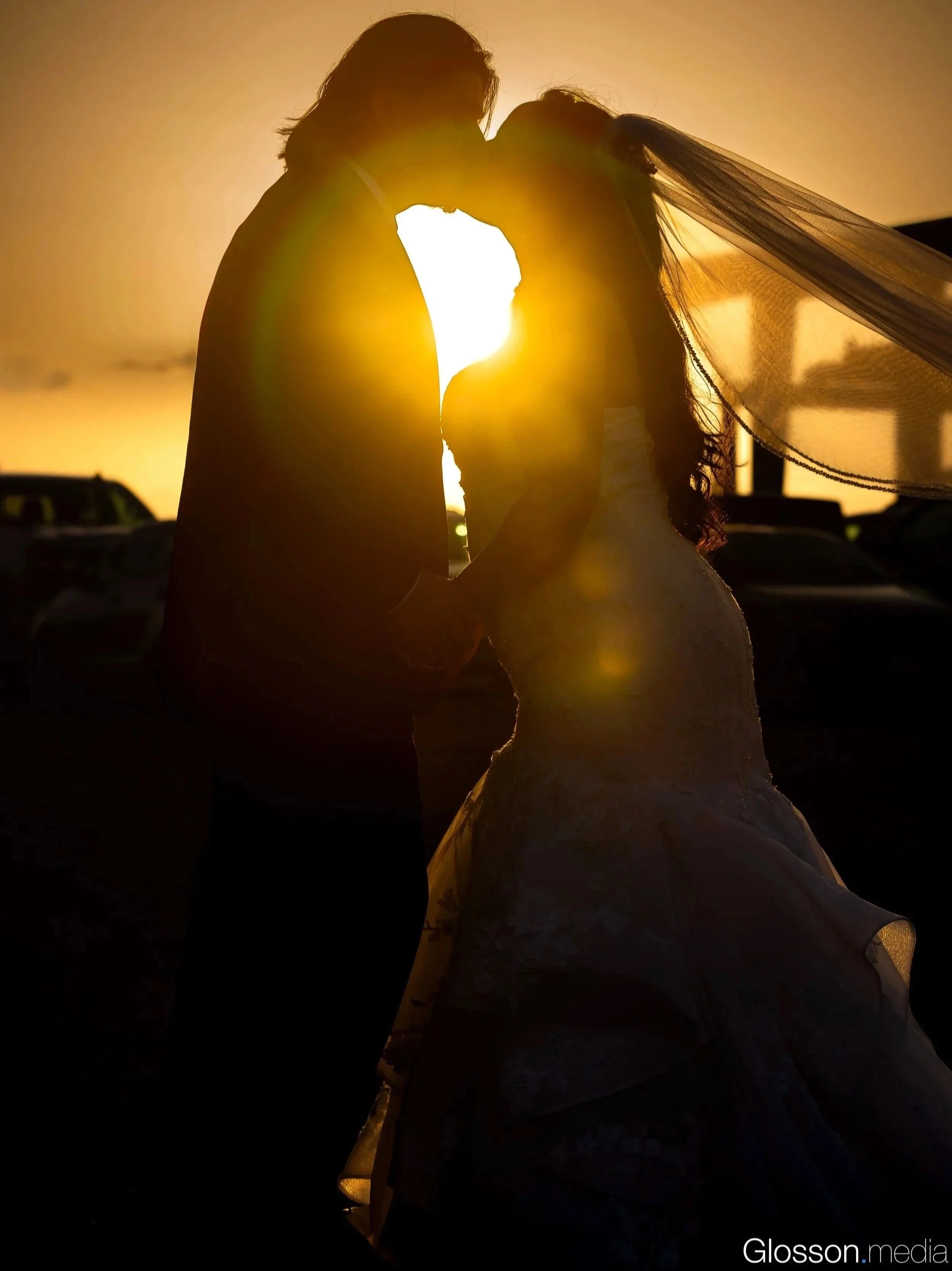 Silhouette of a bride and groom kissing at sunset, holding hands, with cars visible in the background.