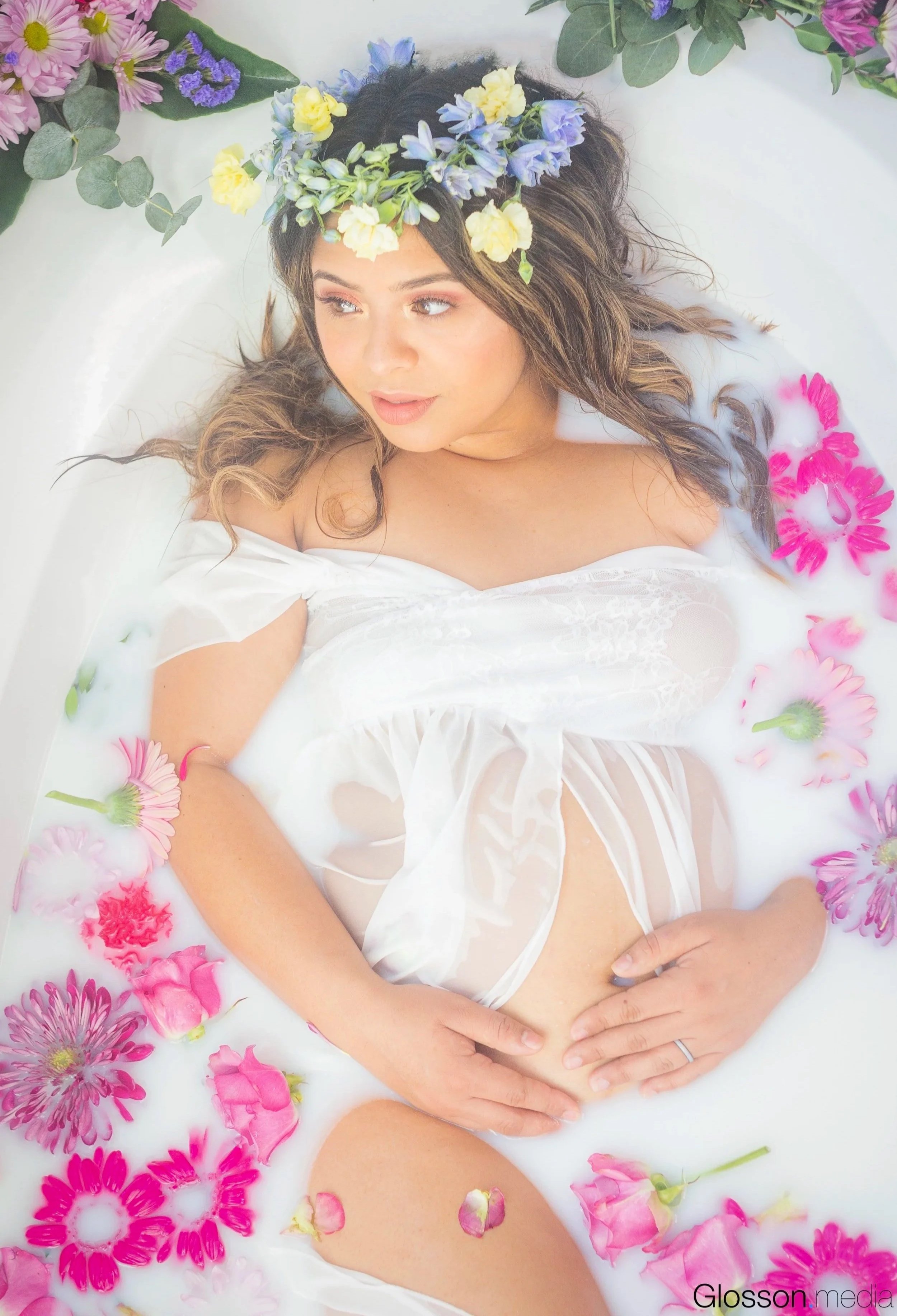 A woman lying in a milk bath with pink and purple flowers surrounding her, wearing a white sheer dress and a floral crown.