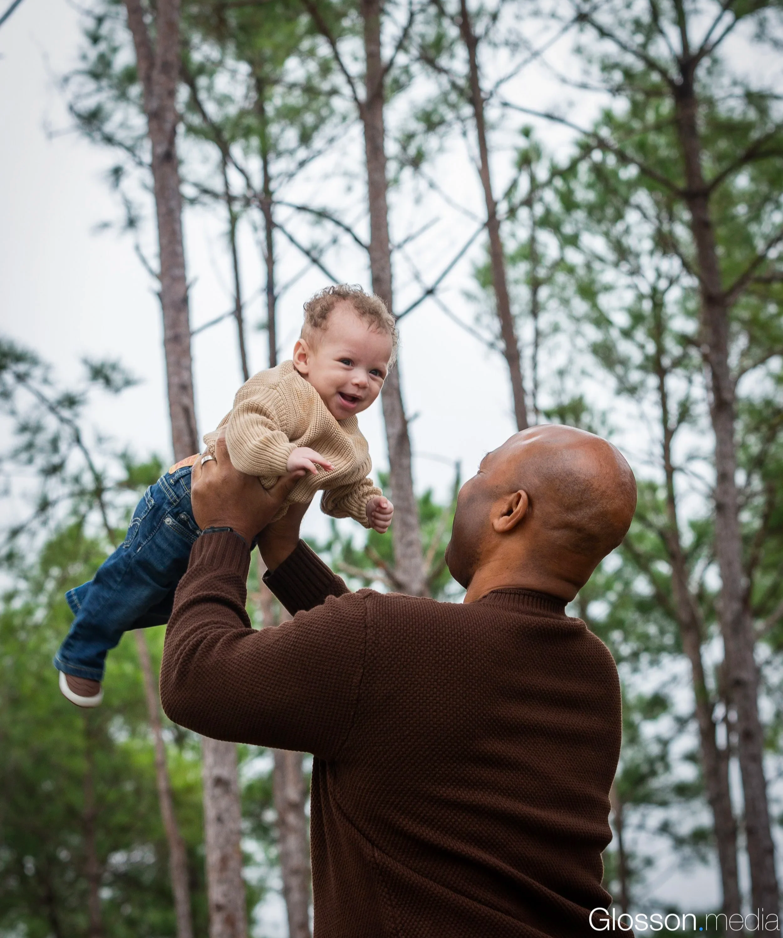 An adult man lifting a happy baby boy in the air outdoors, tall trees in the background.