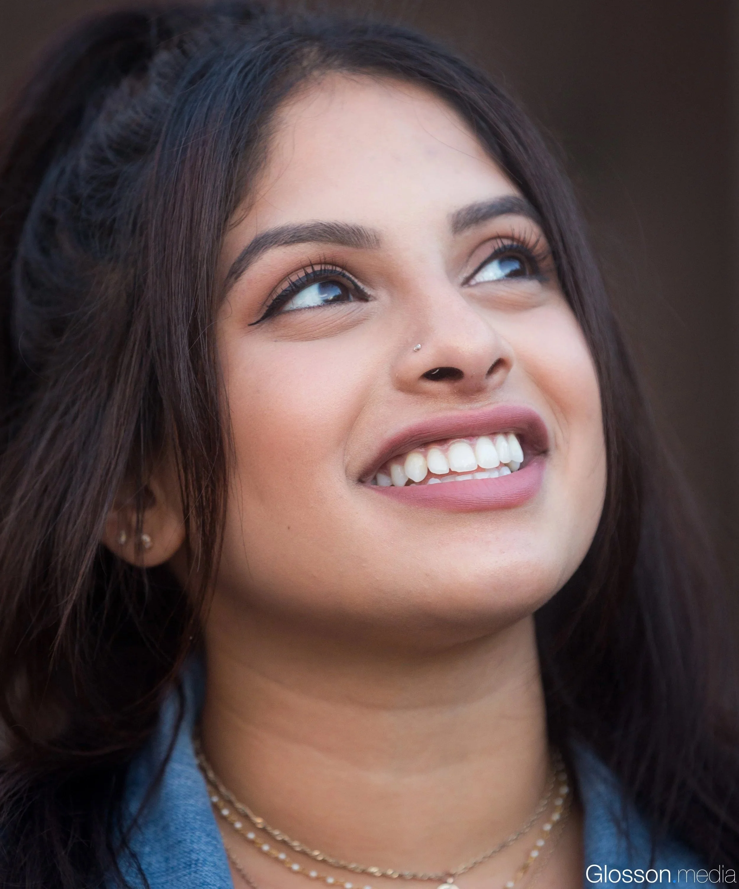 Close-up of a woman's face showing her smiling with white teeth, dark eyebrows, and long eyelashes, wearing layered pearl necklaces, small earrings, and a blue top.