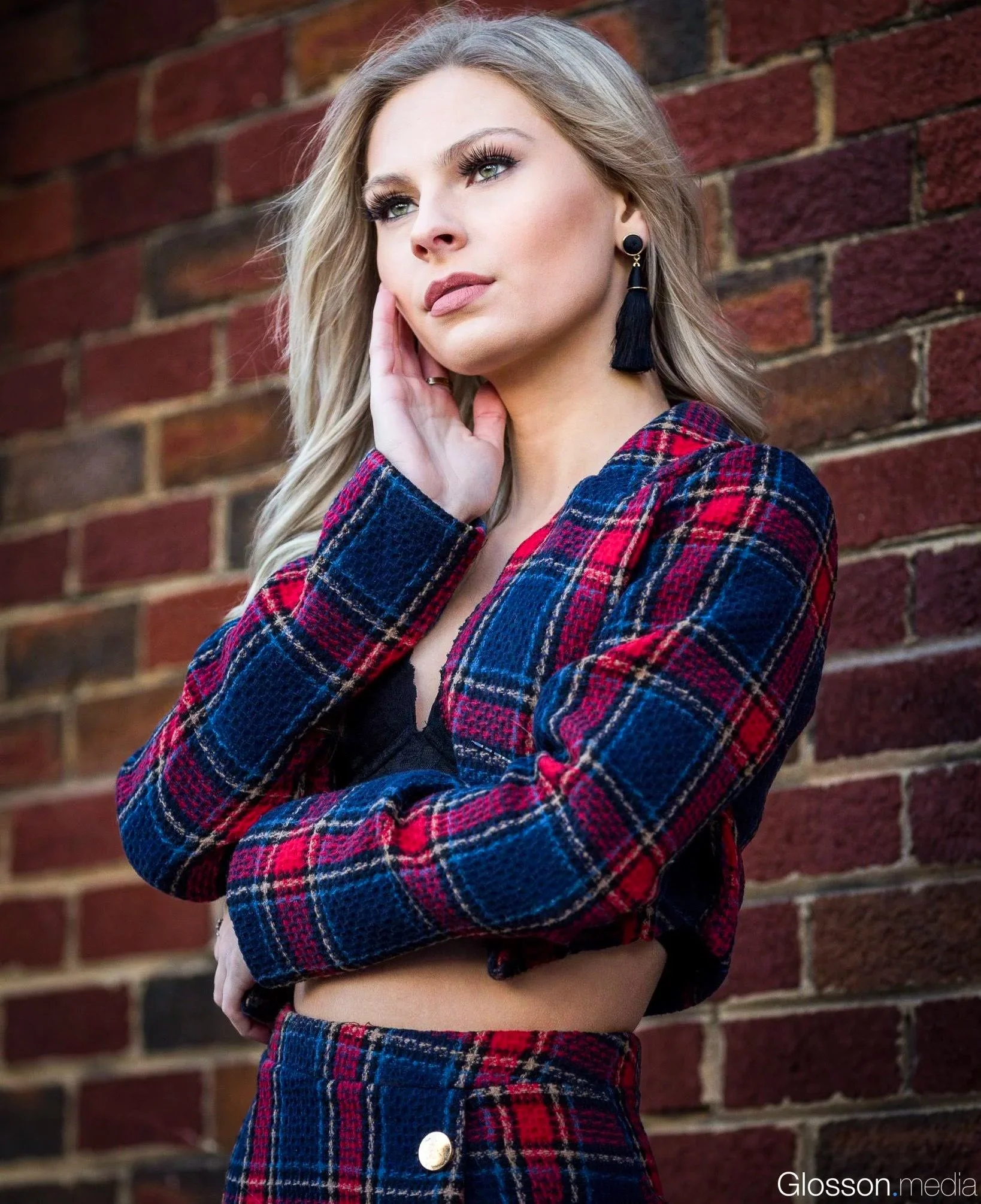 A woman with blonde hair and blue eyes standing against a brick wall, wearing a red and blue plaid jacket and large black tassel earrings.