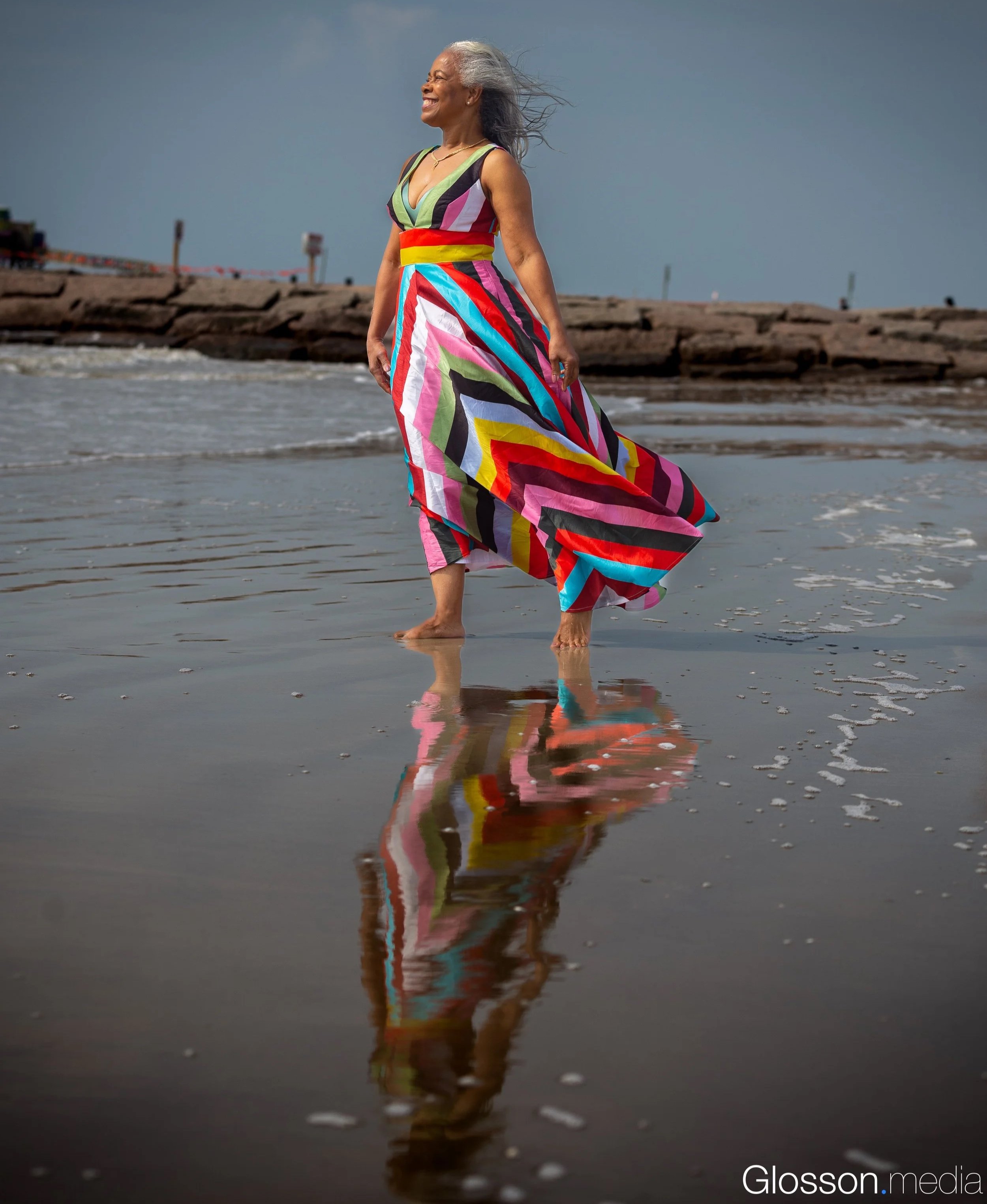 An elderly woman in a colorful patterned dress walking barefoot on the beach with her reflection visible in the shallow water, against a cloudy sky.