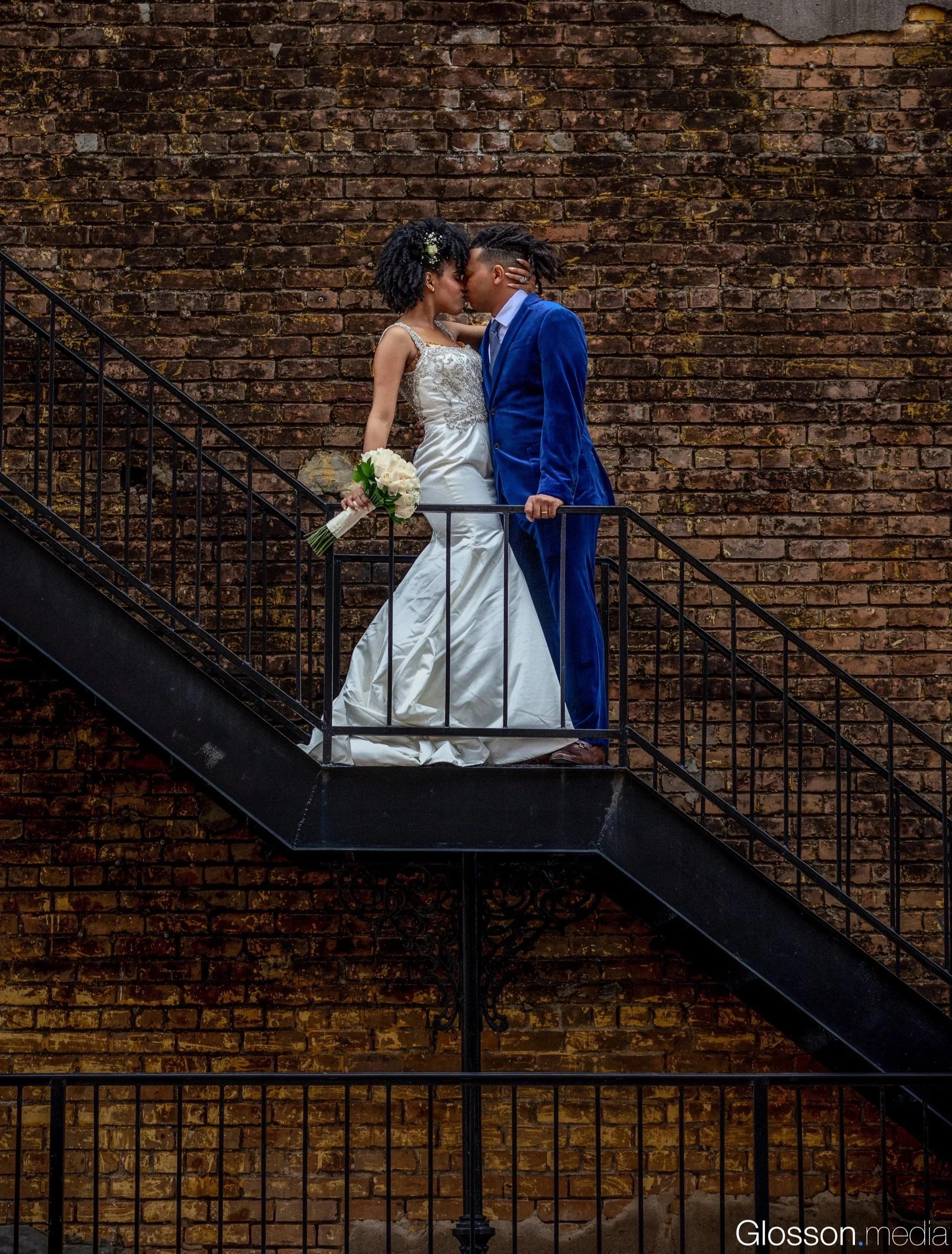 A couple dressed in wedding attire sharing a kiss on a black metal staircase in front of a worn brick wall. The bride is holding a bouquet of white roses.