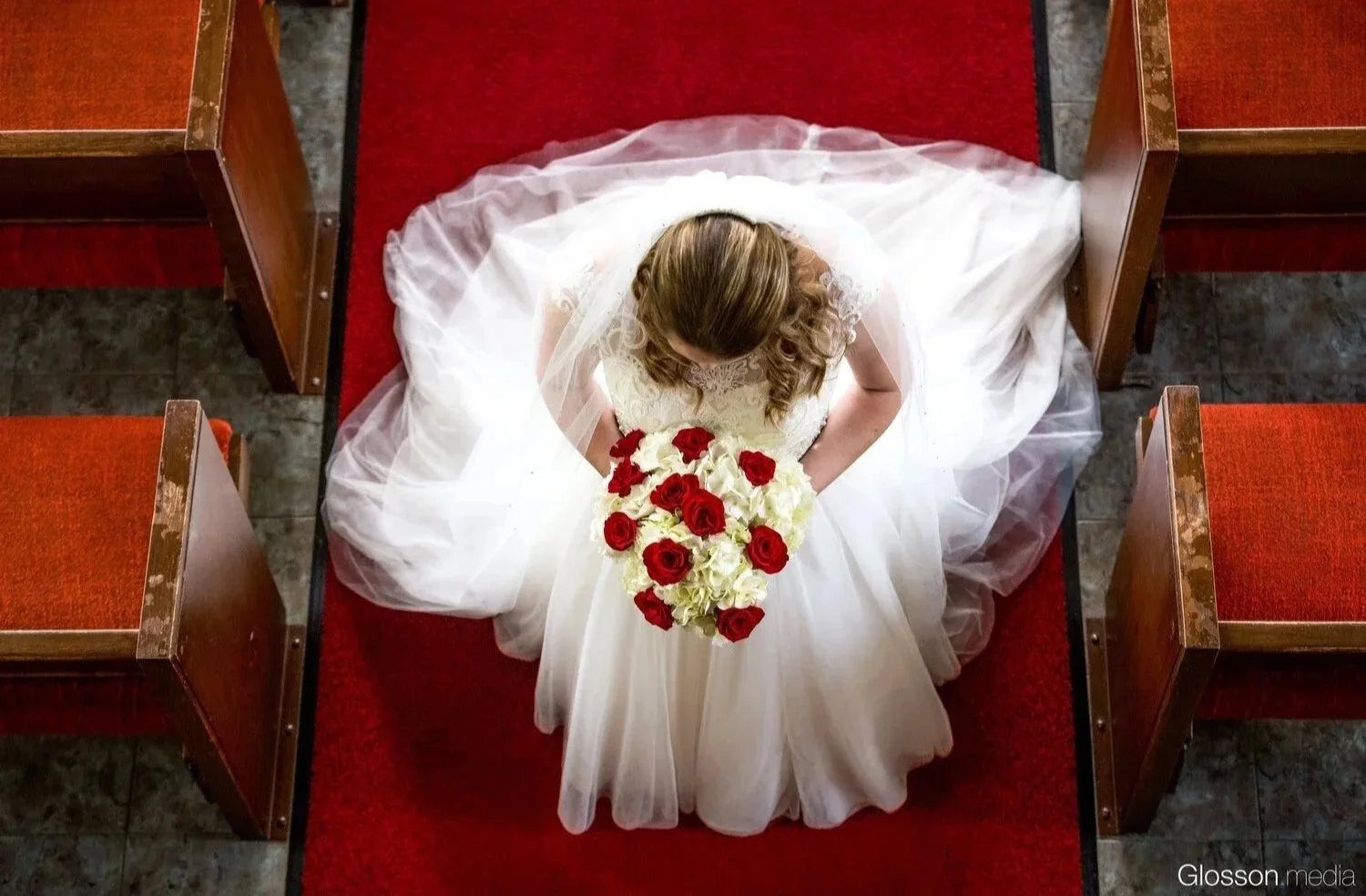 Top-down view of a bride in a white wedding dress sitting on a pew in a church, holding a bouquet of red and white roses, surrounded by wooden pews and a red carpet.