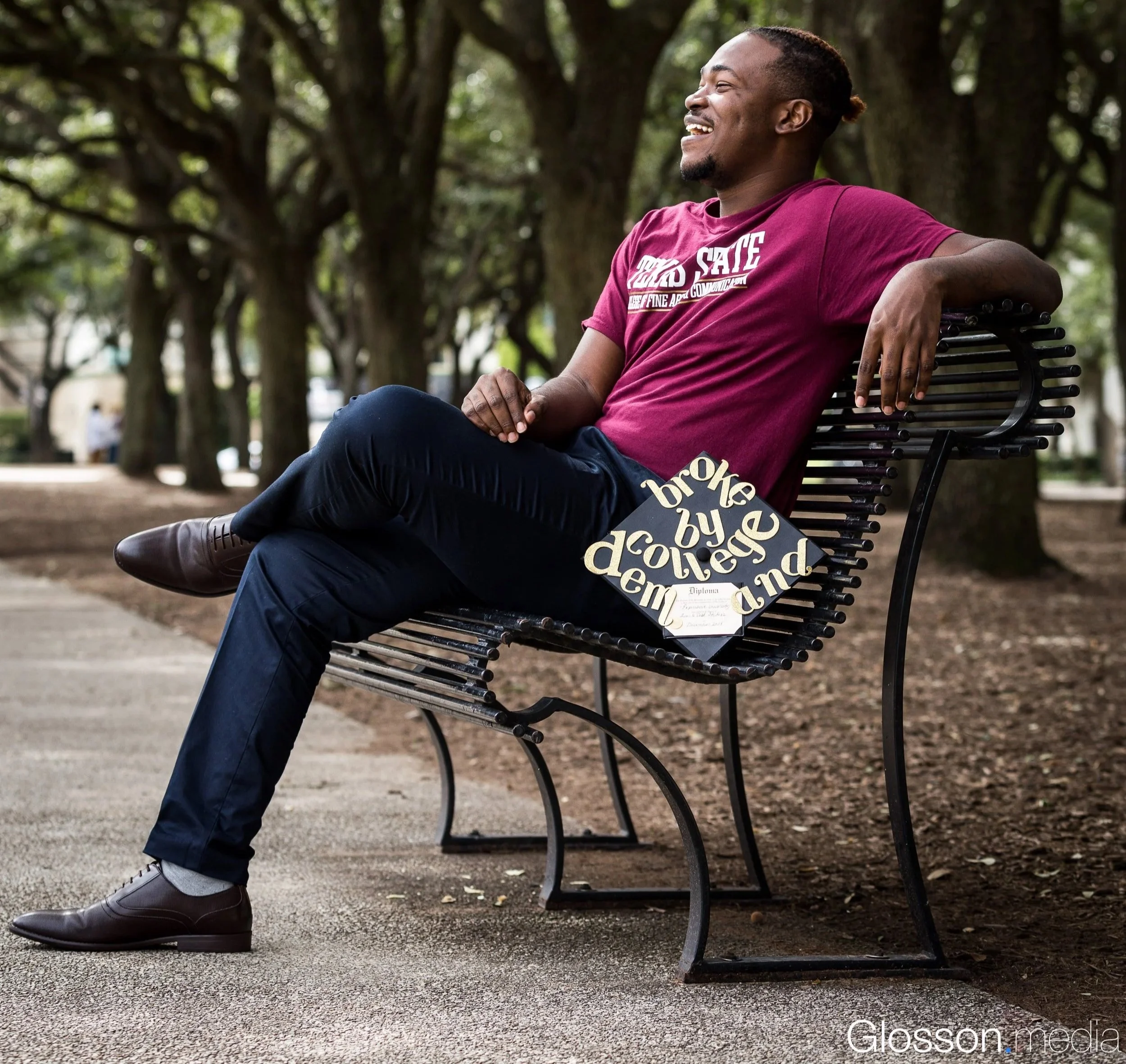 A man sitting on a park bench, smiling and looking relaxed, wearing a maroon T-shirt and dark pants, with a graduation cap with gold lettering resting on his lap, surrounded by trees.