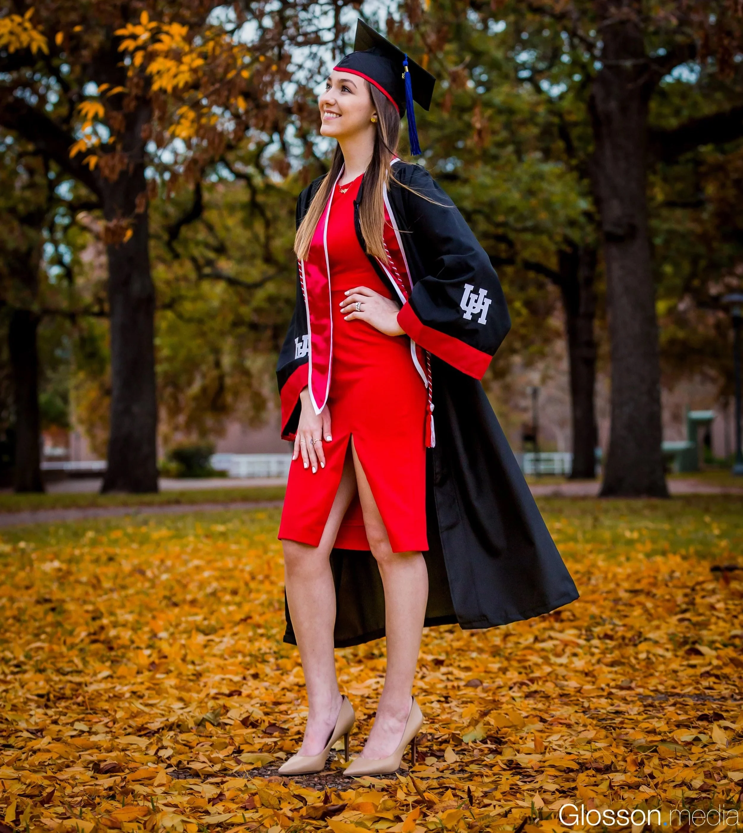 A woman in a red dress and beige high heels celebrating graduation outdoors, wearing a black graduation cap and gown with red accents, standing among autumn leaves with trees in the background.