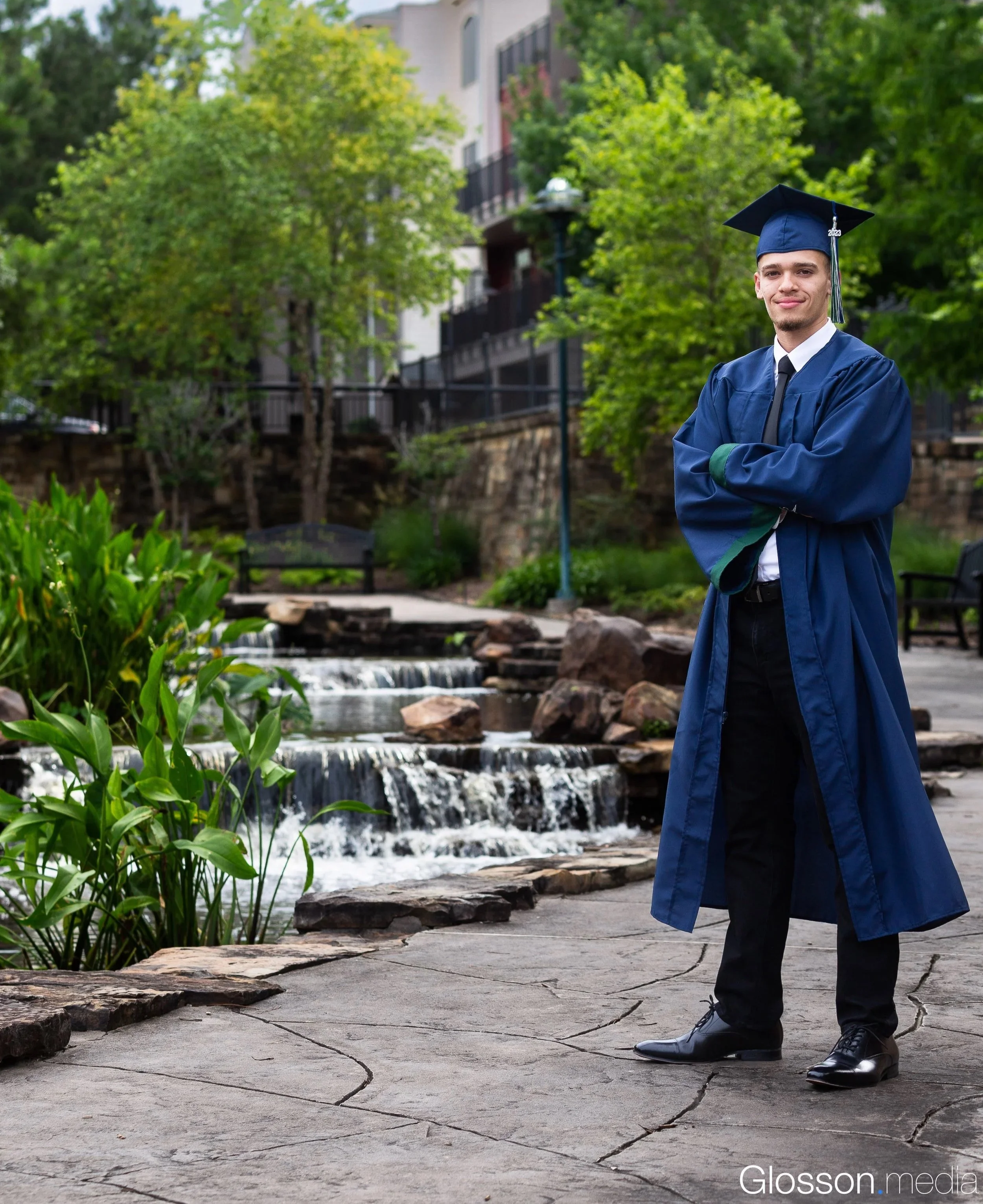 A young man in a cap and gown stands outdoors with arms crossed, smiling, near a small waterfall and lush trees.