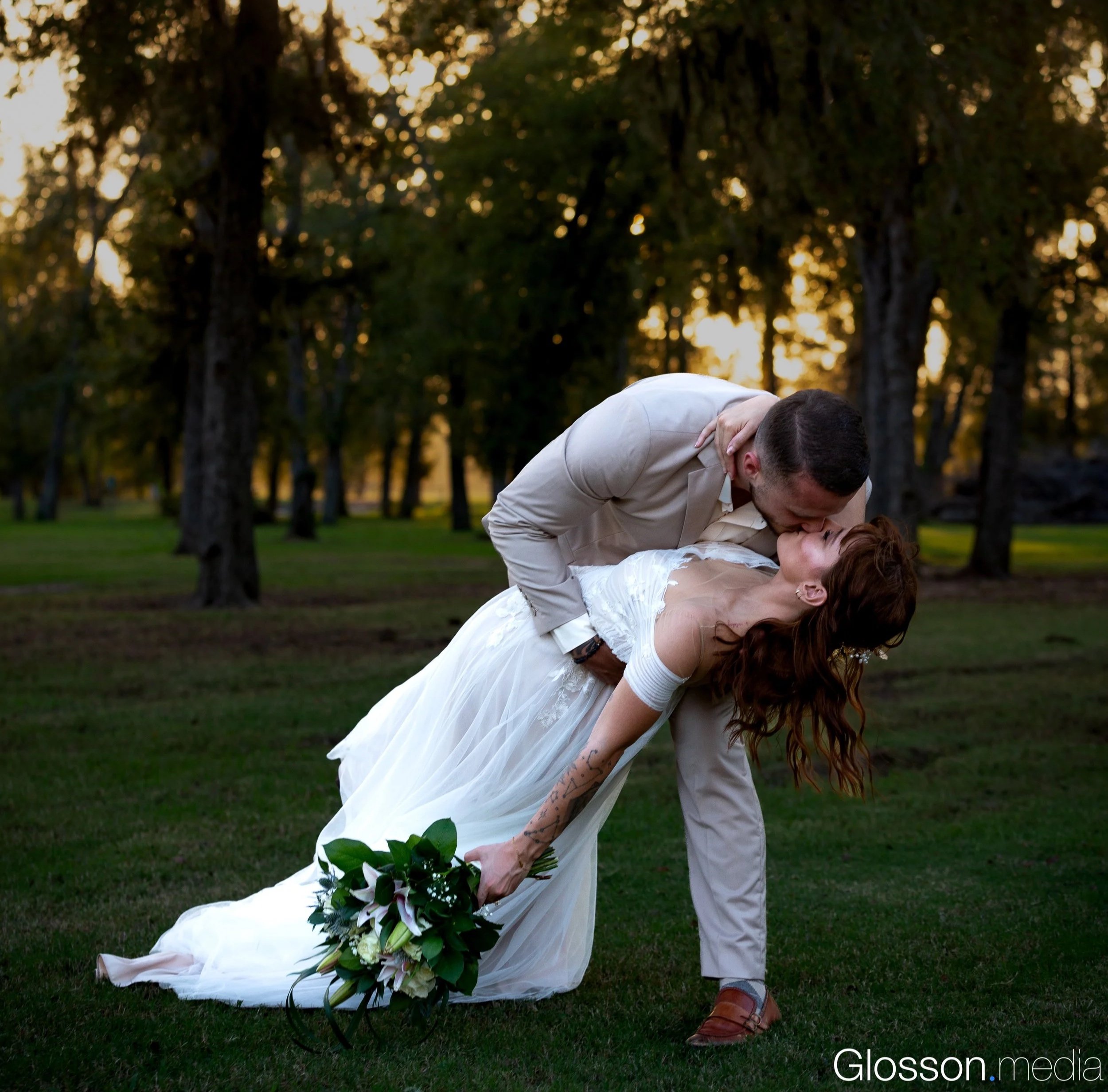 A couple in wedding attire sharing a kiss outdoors at sunset, with trees in the background, the groom leaning forward and the bride holding a bouquet.