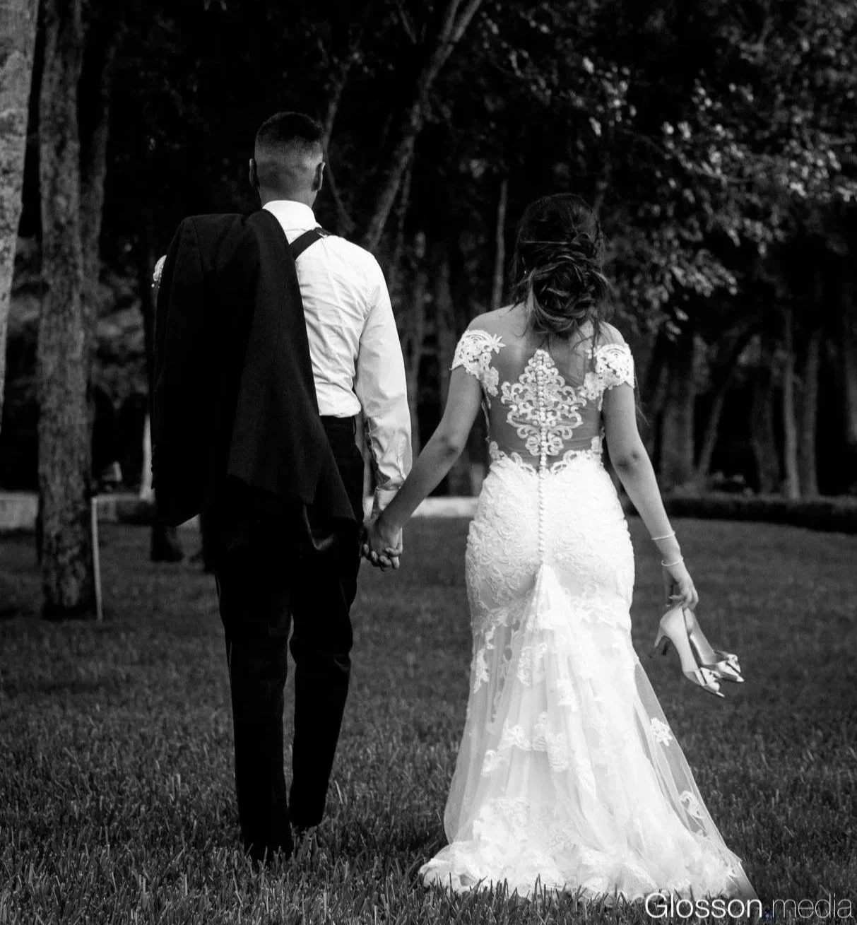 A black and white photo of a bride and groom walking hand in hand outdoors, with the bride holding her high heels in one hand and wearing a wedding dress with lace details and an open back.